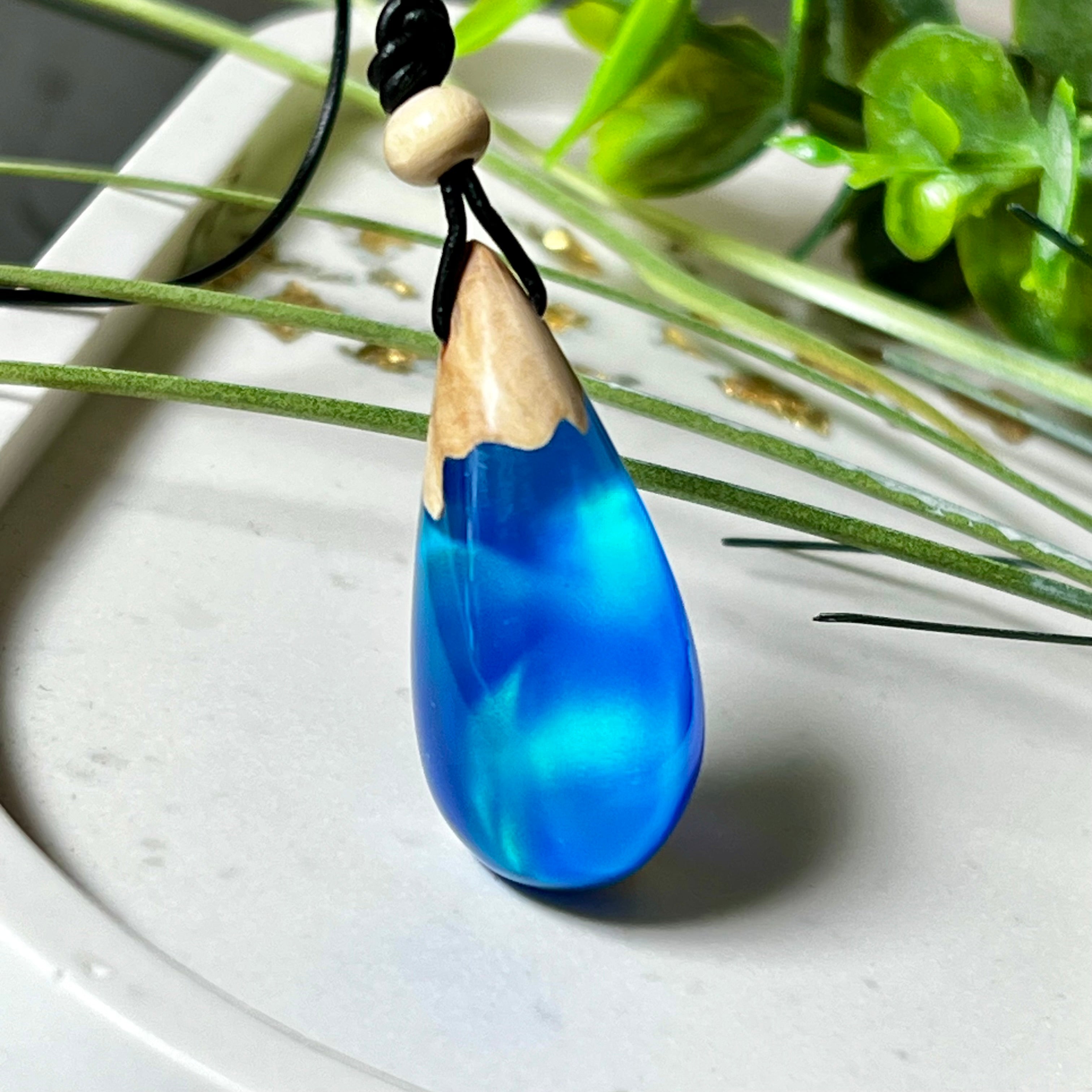 Handgefertigter Tropfenanhänger aus Maserholz und Harz mit synthetischem Opal. Die Farben schimmern je nach Lichteinfall in leuchtendem Blau und Grün – inspiriert von Polarlichtern. Der Anhänger ist ca. 3,9 × 1,8 cm groß und hängt an einem verstellbaren Lederband mit Holzperlen. Jedes Stück ist ein Unikat mit einzigartiger Holzmaserung. Ideal als Geschenk oder stilvoller Begleiter im Alltag.
