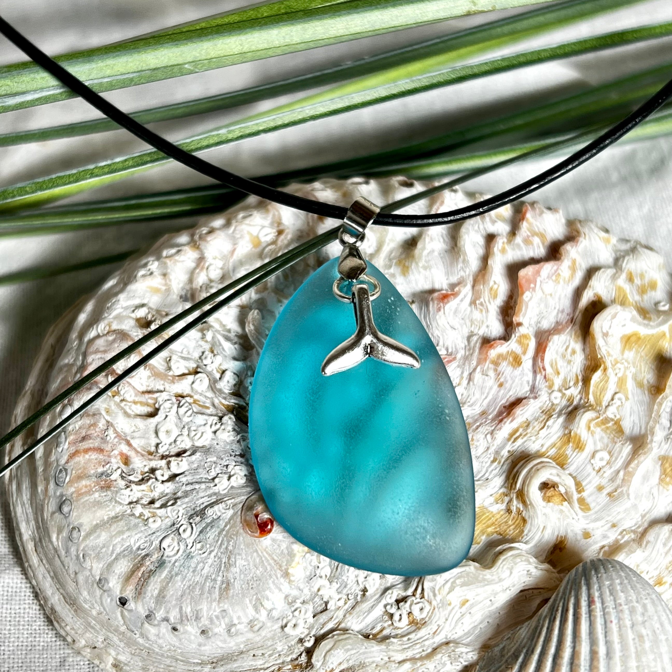 Edle Halskette „Blue Essence“ mit handgefertigtem türkisfarbenem Seeglas-Anhänger und Edelstahl-Flosse. Die Kette besteht aus schwarzer Lederkordel mit Karabinerverschluss und Verlängerung aus Edelstahl. Das matte Seeglas reflektiert Licht wie Meereswasser – ideal für Strandliebhaber und Naturliebhaber.