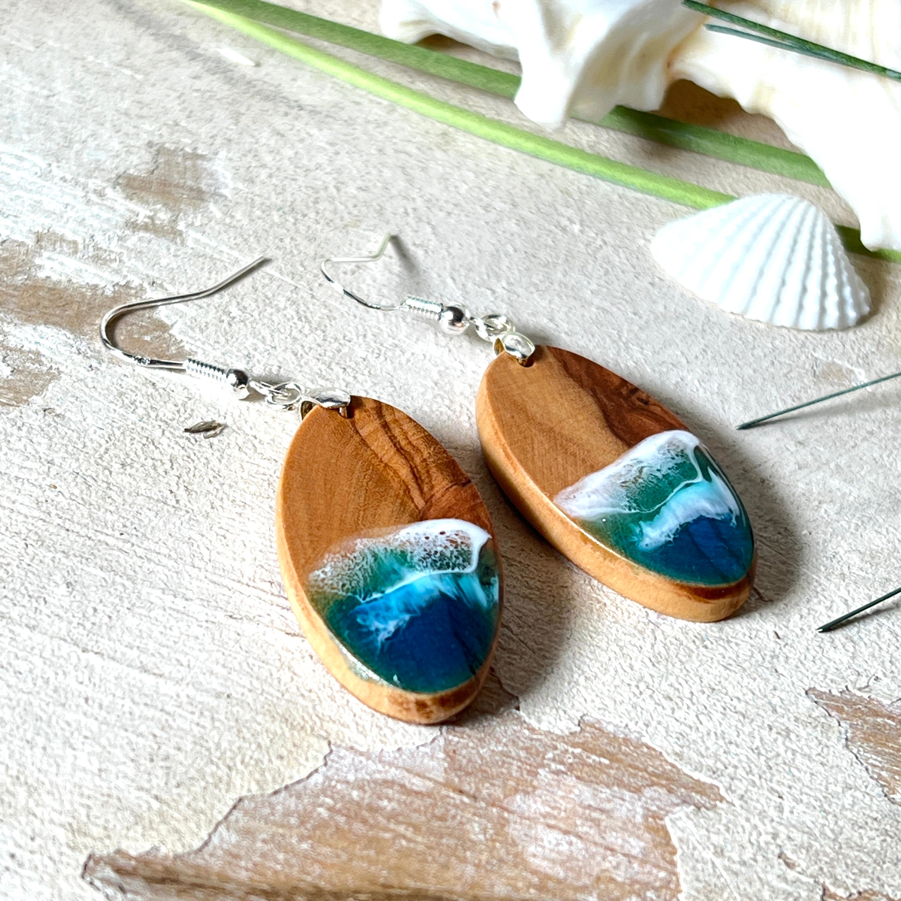 Handgefertigte Ohrringe „Wave Harmony“ aus Olivenholz und Epoxidharz in Blau-, Türkis- und Weißtönen. Das Design erinnert an Meereswellen am Strand. Jeder Ohrring ist ein Unikat mit individueller Maserung. Versilberte, nickelfreie und bleifreie Anhängerschlaufe, 925 Silber Ohrhaken. Maritimer Schmuck – perfekt für Meerliebhaber und als edles Geschenk.