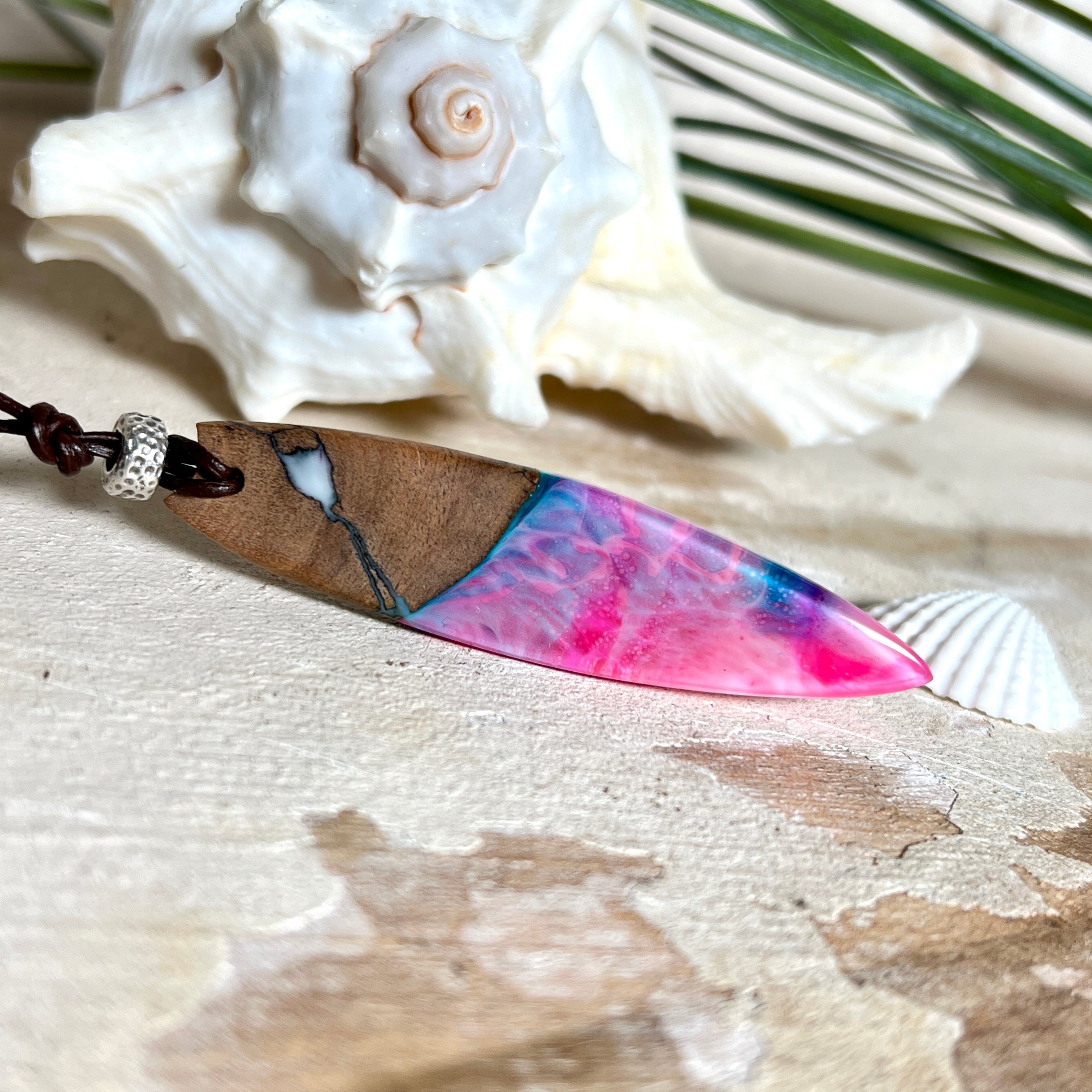 Handgefertigter Surfbrett-Anhänger aus Nussbaumholz und fluoreszierendem Epoxidharz in Pink, Blau und Weiß. Der Anhänger leuchtet zart bei Schwarzlicht und wird an einem verstellbaren Lederband (25–44 cm) mit Holzperlen getragen. Ein maritimes Unikat – ideal für Surfer und Naturliebhaber.