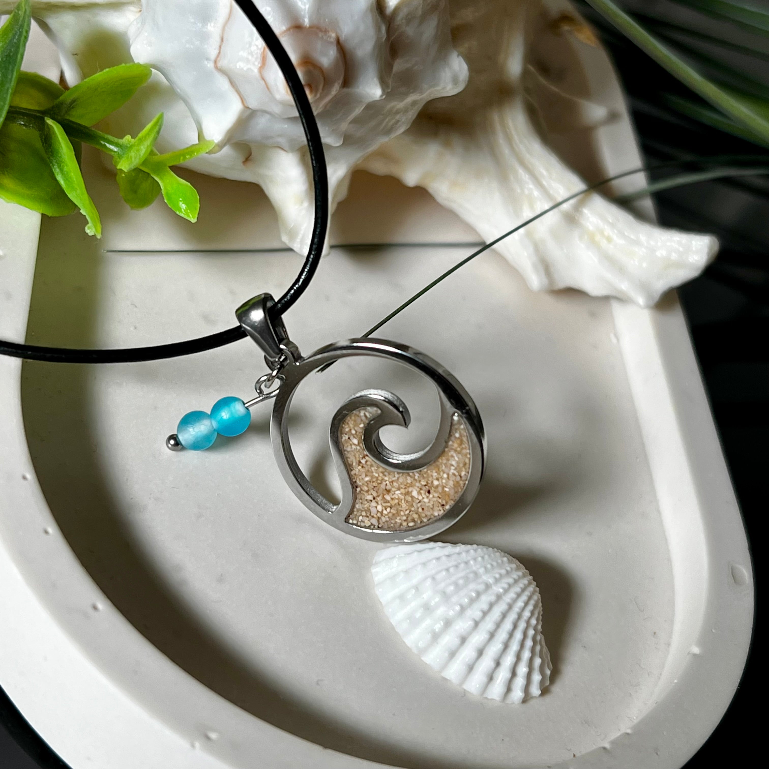Wave Spirit – runder Edelstahlanhänger mit echter Sandfüllung in Harz eingebettet, inspiriert von einer stilisierten Welle. Verziert mit zwei blauen Achatperlen. Der Anhänger (ca. 2,5 cm Durchmesser) hängt an einer 45 cm langen Lederkette mit Edelstahlverschluss und 5 cm Verlängerungskette. Maritimer Look trifft moderne Eleganz – ein handgefertigtes Schmuckstück für Meerliebhaber