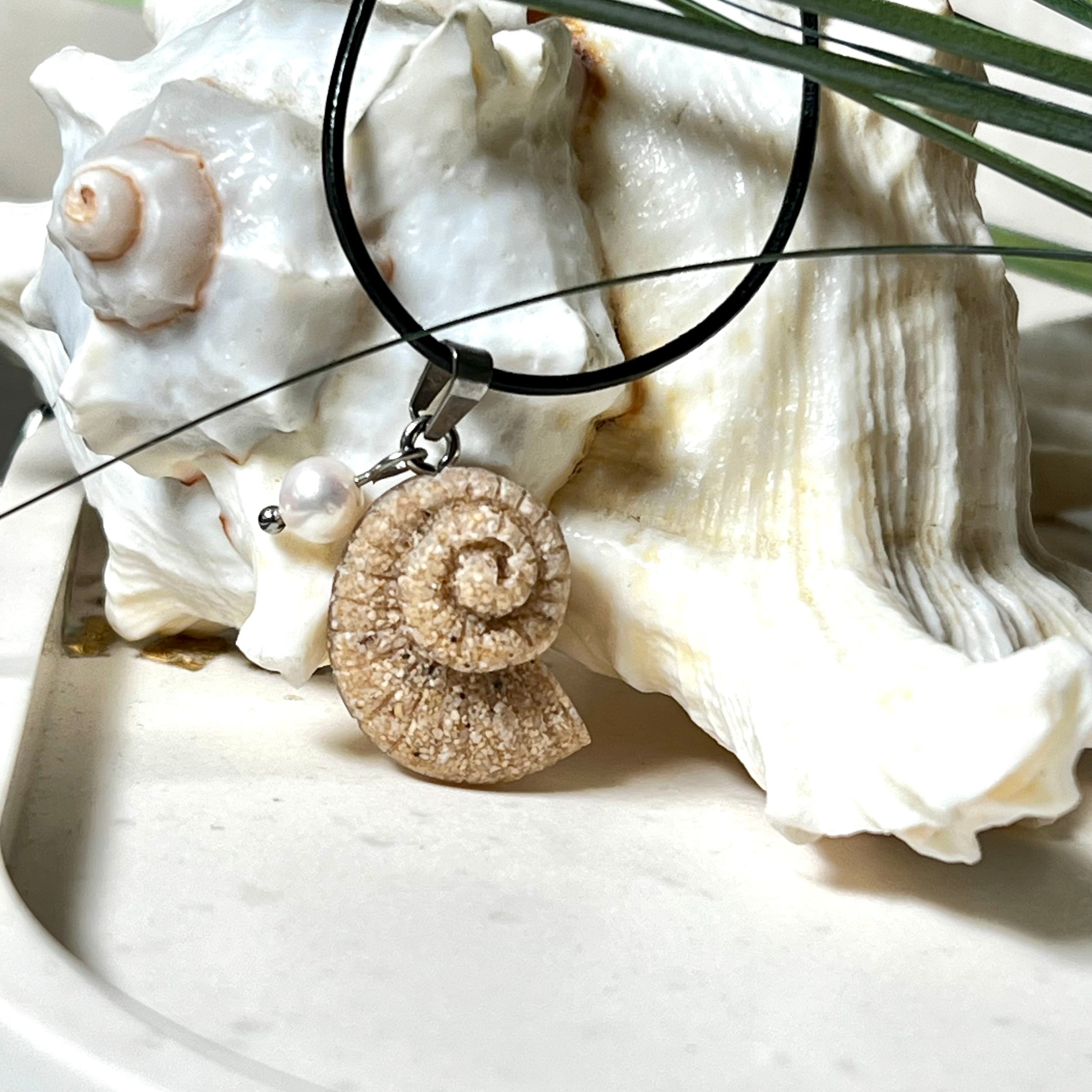 Handgefertigter Anhänger in Ammonit-Form aus echtem Meersand und Harz, kombiniert mit einer weißen Süßwasserperle. Der maritime Anhänger hängt an einer schwarzen Lederkette mit Edelstahlöse und Karabinerverschluss. Im Hintergrund liegt eine weiße Muschel, die das natürliche, strandinspirierte Design des Schmuckstücks unterstreicht. Ideal als besonderes Geschenk oder Erinnerung an den Sommer.