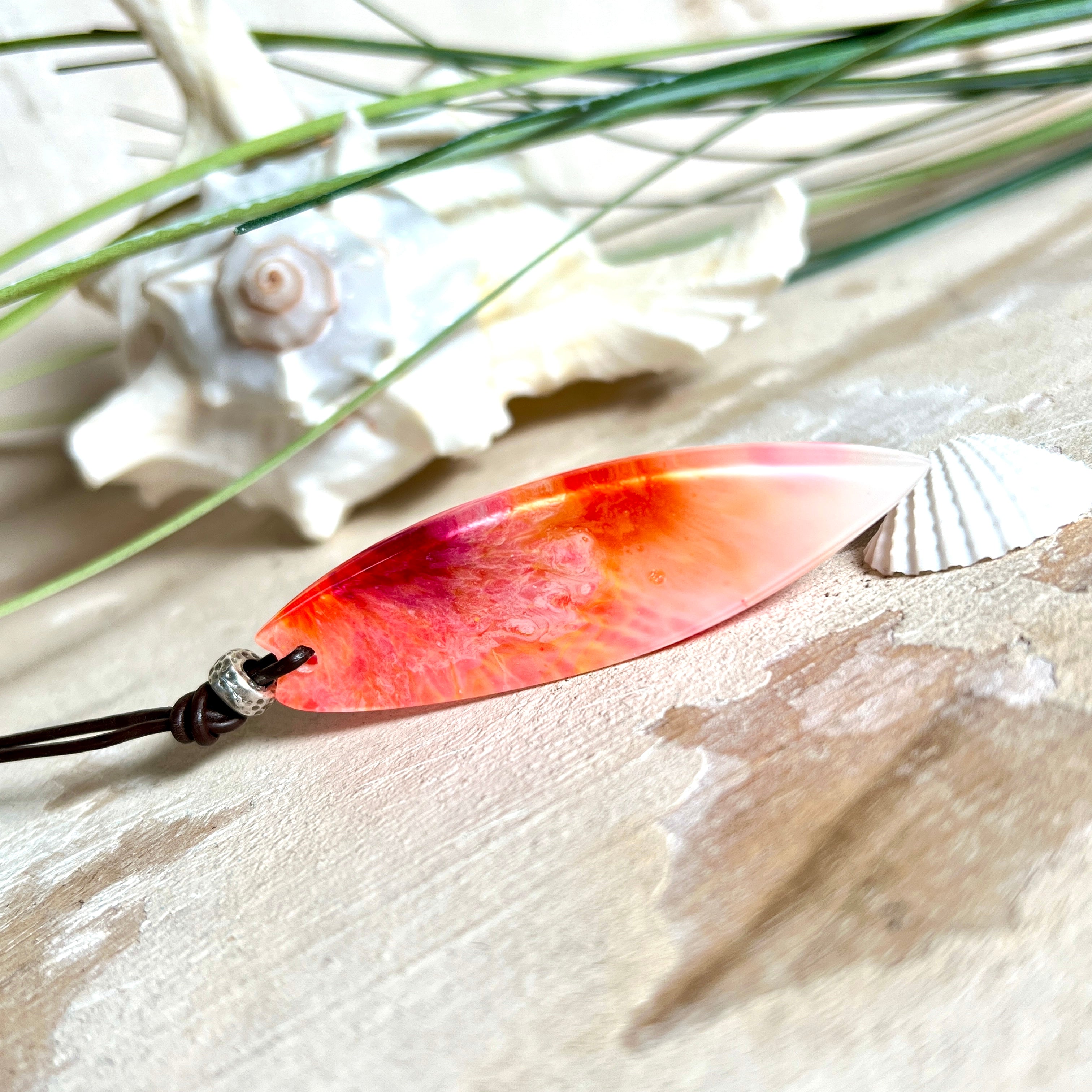 Handgefertigter Surfbrett-Anhänger aus fluoreszierendem Epoxidharz in leuchtenden Orange-, Rot- und Gelbtönen. Leuchtet bei Schwarzlicht. Kombiniert mit verstellbarem Lederband (26–45 cm), verziert mit Spacer- und Holzperlen. Das Design ist inspiriert von der Farbintensität eines Sonnenuntergangs über dem Meer.