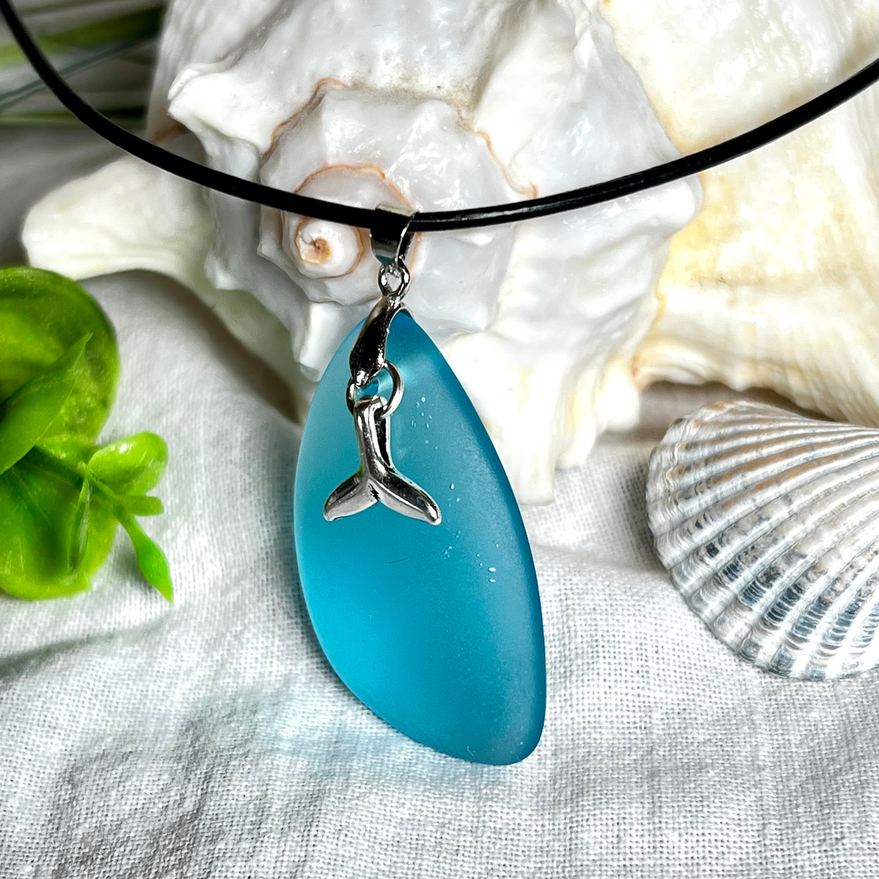Edle Halskette „Blue Essence“ mit handgefertigtem türkisfarbenem Seeglas-Anhänger und Edelstahl-Flosse. Die Kette besteht aus schwarzer Lederkordel mit Karabinerverschluss und Verlängerung aus Edelstahl. Das matte Seeglas reflektiert Licht wie Meereswasser – ideal für Strandliebhaber und Naturliebhaber.