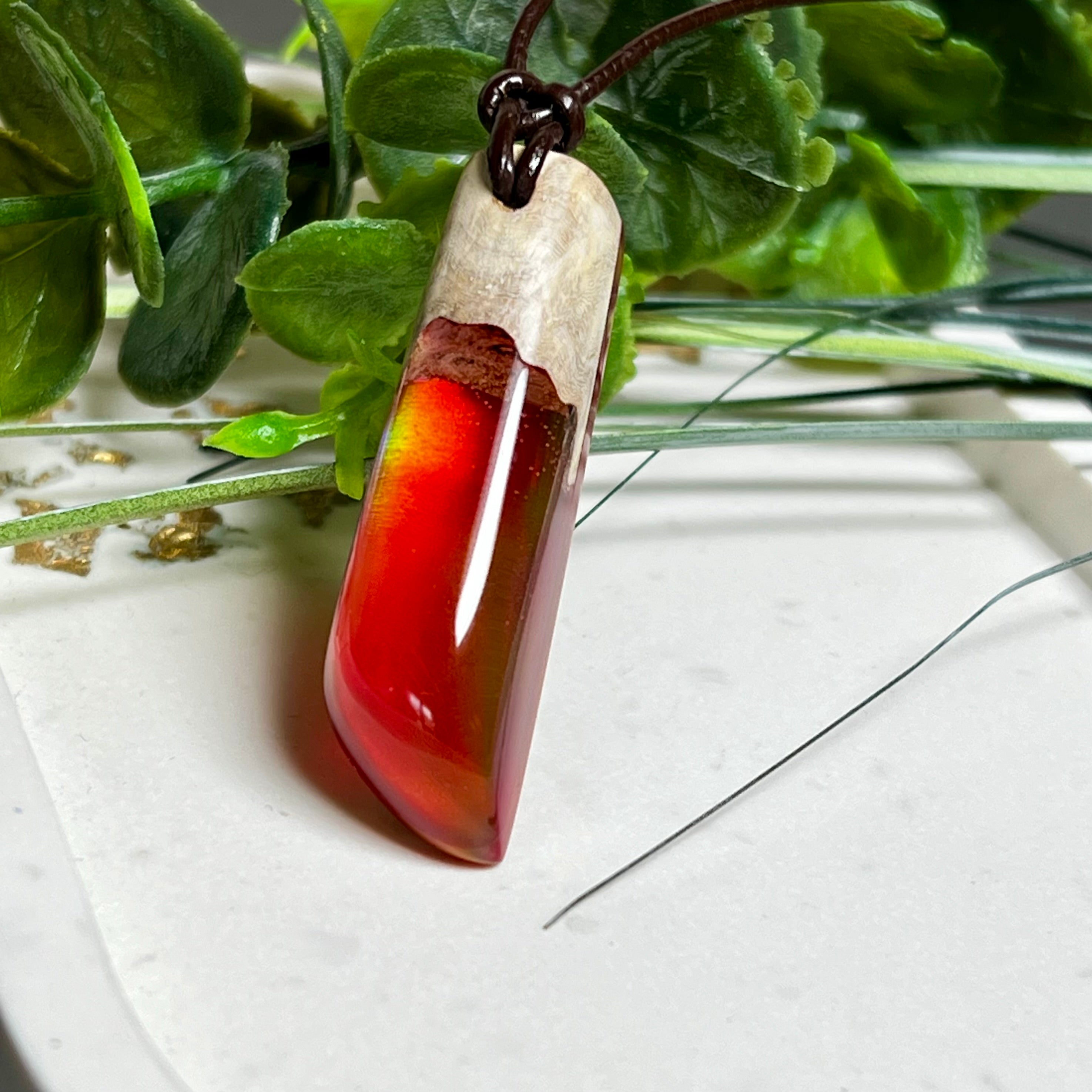 Handgefertigter Hei Toki Anhänger aus Goldfield-Maserholz und rubinrotem Labor-Opal, eingebettet in glänzendes Epoxidharz. Je nach Lichteinfall schimmert das Harz in warmen Farben mit Regenbogeneffekt. Der Anhänger ist ca. 4,3 × 1,5 cm groß und wird an einem verstellbaren Lederband mit Holzperlen getragen. Symbolischer Unikatschmuck – ideal als Geschenk für besondere Menschen.