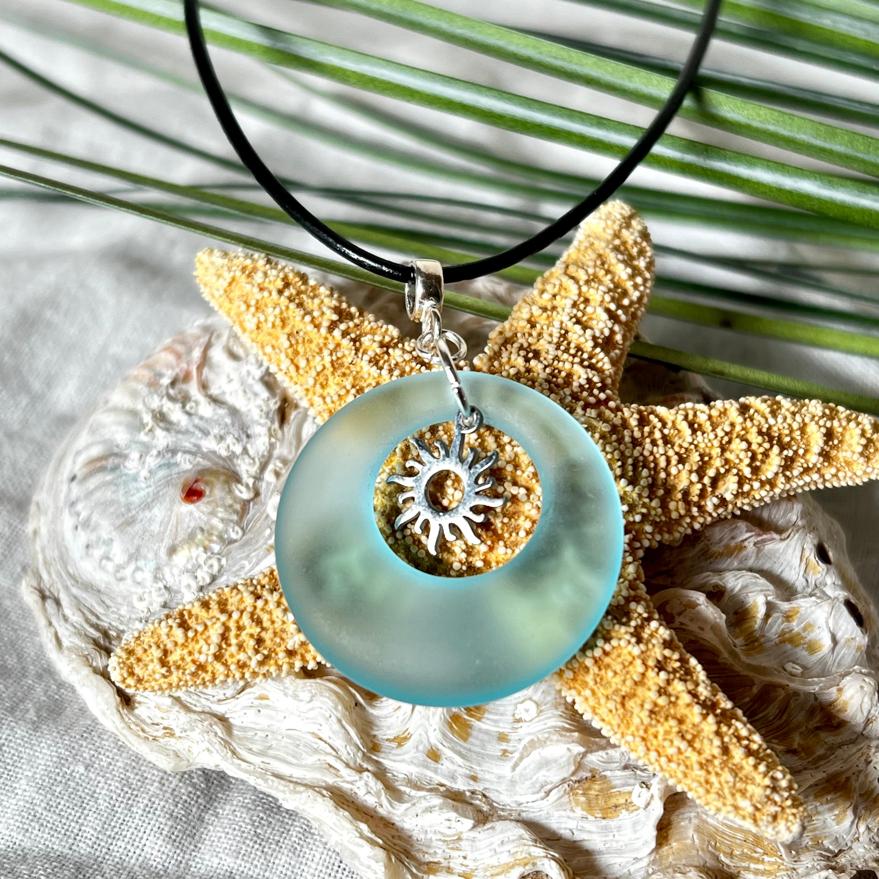 Handgefertigte Halskette mit Donut-Anhänger aus türkisfarbenem Seeglas, kombiniert mit einem filigranen Sonnen-Charm aus Edelstahl. Die Kette besteht aus einer schwarzen Lederschnur (1,5 mm) mit Karabiner und Verlängerungskette aus Edelstahl. Maritimes Schmuckstück für Meerliebhaberinnen und Sommerfreunde. Unikat mit stilvoller Ausstrahlung.