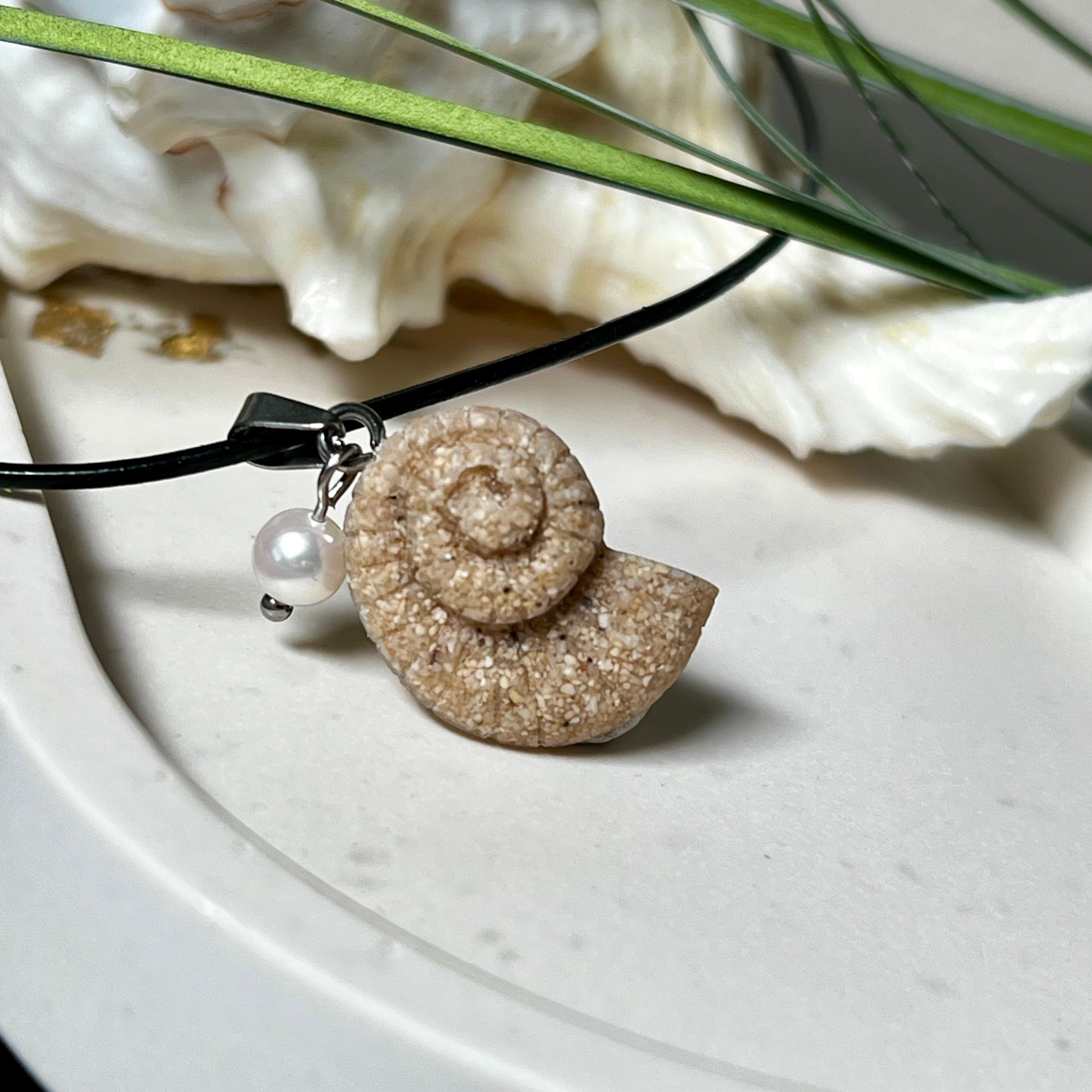 Handgefertigter Anhänger in Ammonit-Form aus echtem Meersand und Harz, kombiniert mit einer weißen Süßwasserperle. Der maritime Anhänger hängt an einer schwarzen Lederkette mit Edelstahlöse und Karabinerverschluss. Im Hintergrund liegt eine weiße Muschel, die das natürliche, strandinspirierte Design des Schmuckstücks unterstreicht. Ideal als besonderes Geschenk oder Erinnerung an den Sommer.