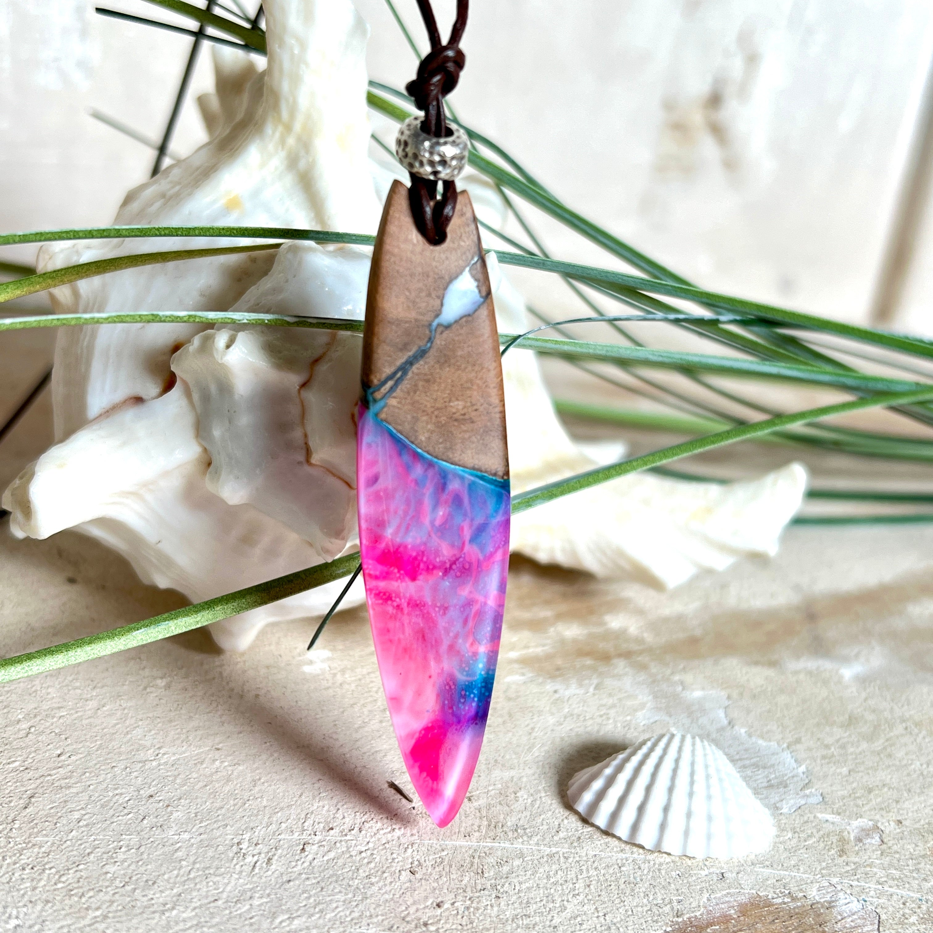 Handgefertigter Surfbrett-Anhänger aus Nussbaumholz und fluoreszierendem Epoxidharz in Pink, Blau und Weiß. Der Anhänger leuchtet zart bei Schwarzlicht und wird an einem verstellbaren Lederband (25–44 cm) mit Holzperlen getragen. Ein maritimes Unikat – ideal für Surfer und Naturliebhaber.