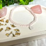 Handgefertigtes Rosenquarz-Armband mit facettierten 4mm Perlen, ergänzt durch einen filigranen Blume-des-Lebens-Anhänger aus 925er Silber. Präsentiert auf hellem Hintergrund mit Rosenquarzsteinen und goldenen Dekoelementen. Spiritueller Schmuck mit harmonisierender Wirkung.