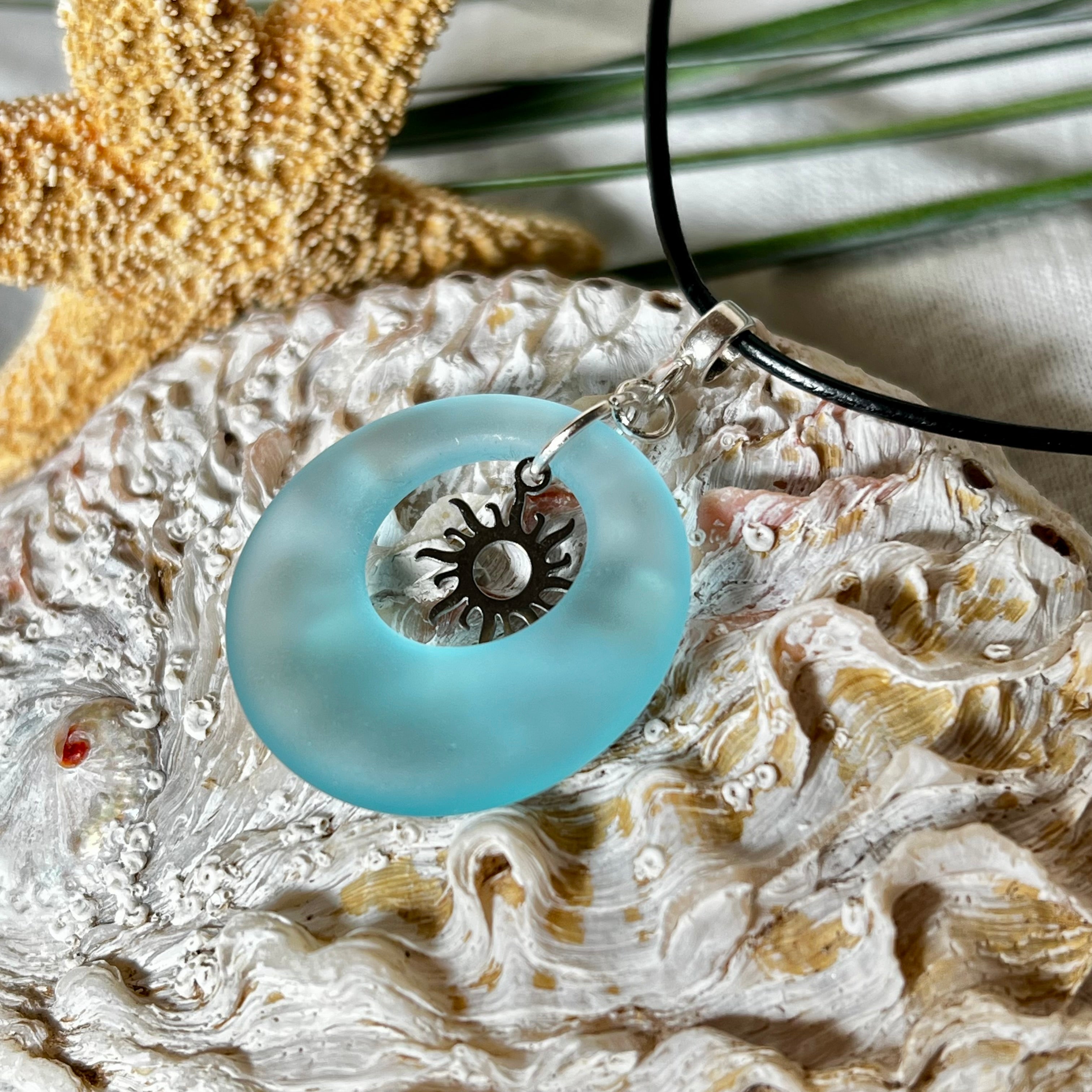 Handgefertigte Halskette mit Donut-Anhänger aus türkisfarbenem Seeglas, kombiniert mit einem filigranen Sonnen-Charm aus Edelstahl. Die Kette besteht aus einer schwarzen Lederschnur (1,5 mm) mit Karabiner und Verlängerungskette aus Edelstahl. Maritimes Schmuckstück für Meerliebhaberinnen und Sommerfreunde. Unikat mit stilvoller Ausstrahlung.