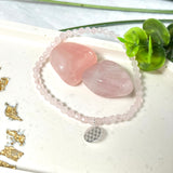 Handgefertigtes Rosenquarz-Armband mit facettierten 4mm Perlen, ergänzt durch einen filigranen Blume-des-Lebens-Anhänger aus 925er Silber. Präsentiert auf hellem Hintergrund mit Rosenquarzsteinen und goldenen Dekoelementen. Spiritueller Schmuck mit harmonisierender Wirkung.