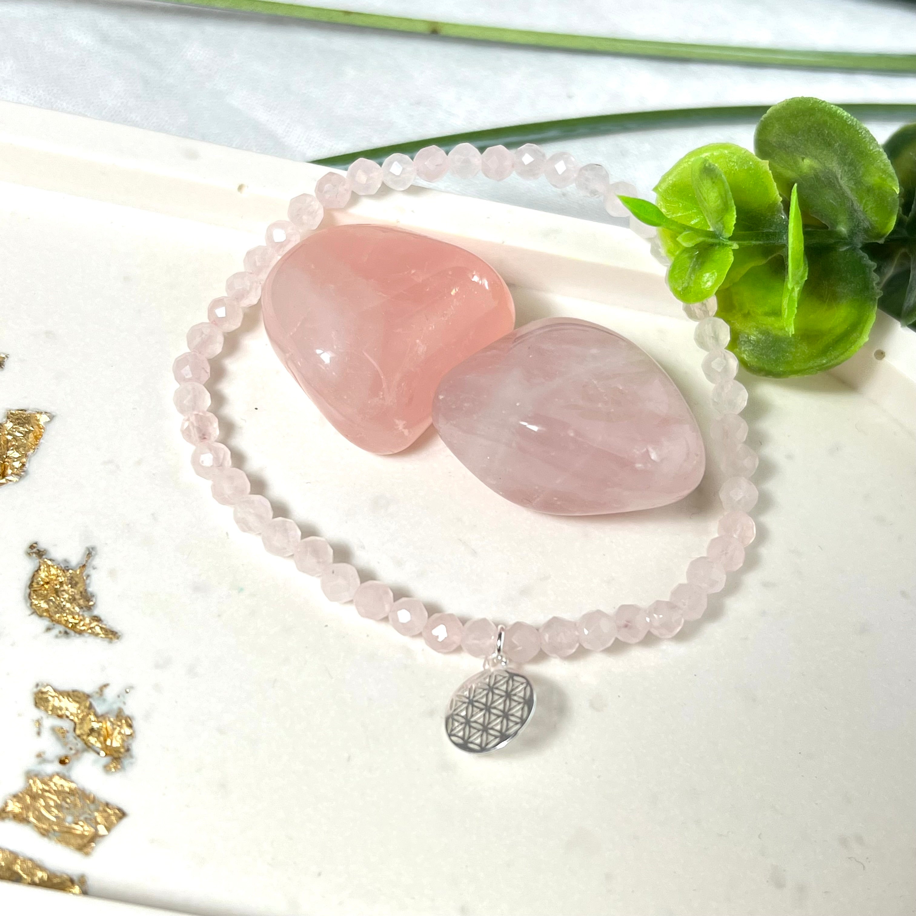 Handgefertigtes Rosenquarz-Armband mit facettierten 4mm Perlen, ergänzt durch einen filigranen Blume-des-Lebens-Anhänger aus 925er Silber. Präsentiert auf hellem Hintergrund mit Rosenquarzsteinen und goldenen Dekoelementen. Spiritueller Schmuck mit harmonisierender Wirkung.