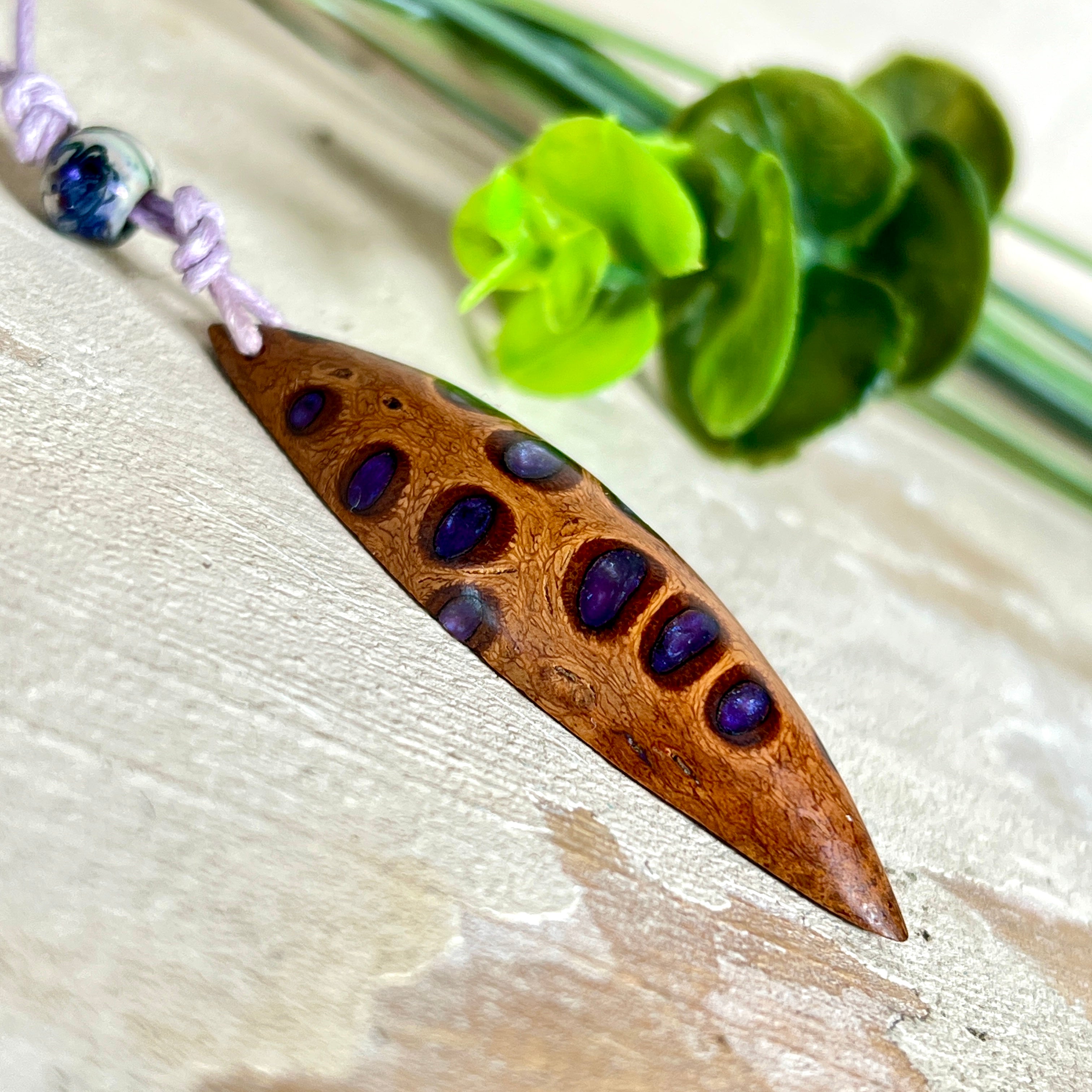 Handgefertigter Anhänger aus einem australischen Banksiazapfen mit eingelassenem metallic-violettem Harz. Der ovale, tropfenförmige Anhänger ist ca. 5,5 cm lang und wird mit einem verstellbaren Baumwollband geliefert. Jedes Stück zeigt die einzigartige natürliche Struktur des Zapfens und schimmert in satten Lilatönen – ein stilvoller Schmuck mit organischem Charakter.