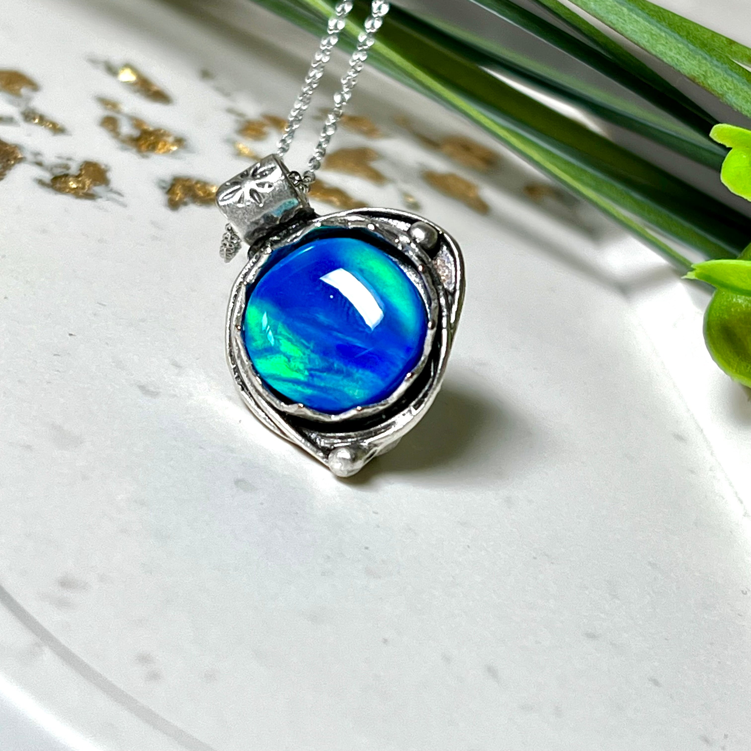 Handgemachter Anhänger mit rundem Cabochon aus Epoxidharz und schimmerndem synthetischem Opal. Die Farben changieren je nach Lichteinfall in Blau, Türkis und Grün. Der Opal sitzt in einer antik versilberten Fassung aus Messing (blei- & nickelfrei). Geliefert mit einer rhodinierten 925 Silberkette (45 cm). Jedes Stück ein Einzelstück – perfekt als stilvolles Geschenk oder persönlicher Glücksbringer.