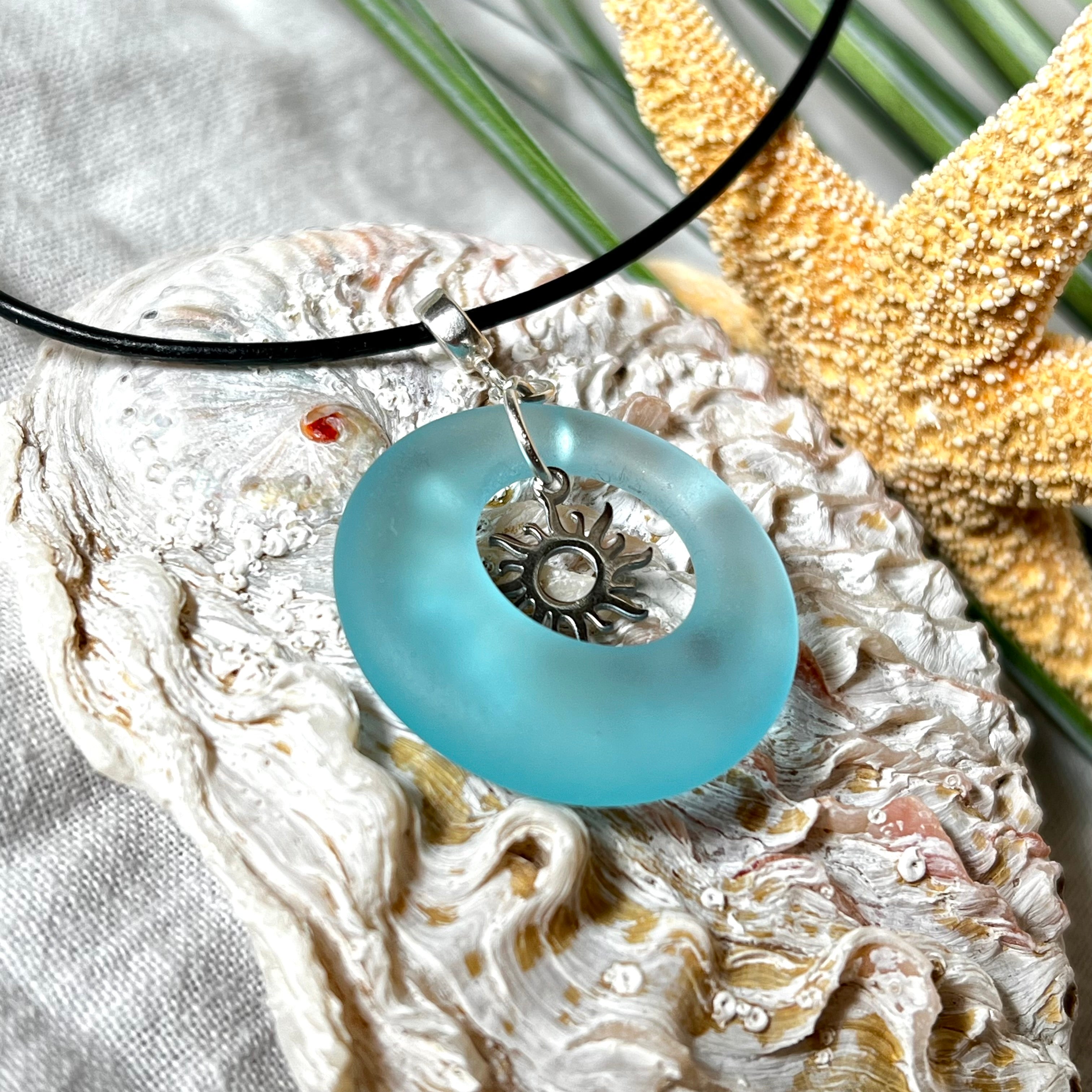 Handgefertigte Halskette mit Donut-Anhänger aus türkisfarbenem Seeglas, kombiniert mit einem filigranen Sonnen-Charm aus Edelstahl. Die Kette besteht aus einer schwarzen Lederschnur (1,5 mm) mit Karabiner und Verlängerungskette aus Edelstahl. Maritimes Schmuckstück für Meerliebhaberinnen und Sommerfreunde. Unikat mit stilvoller Ausstrahlung.
