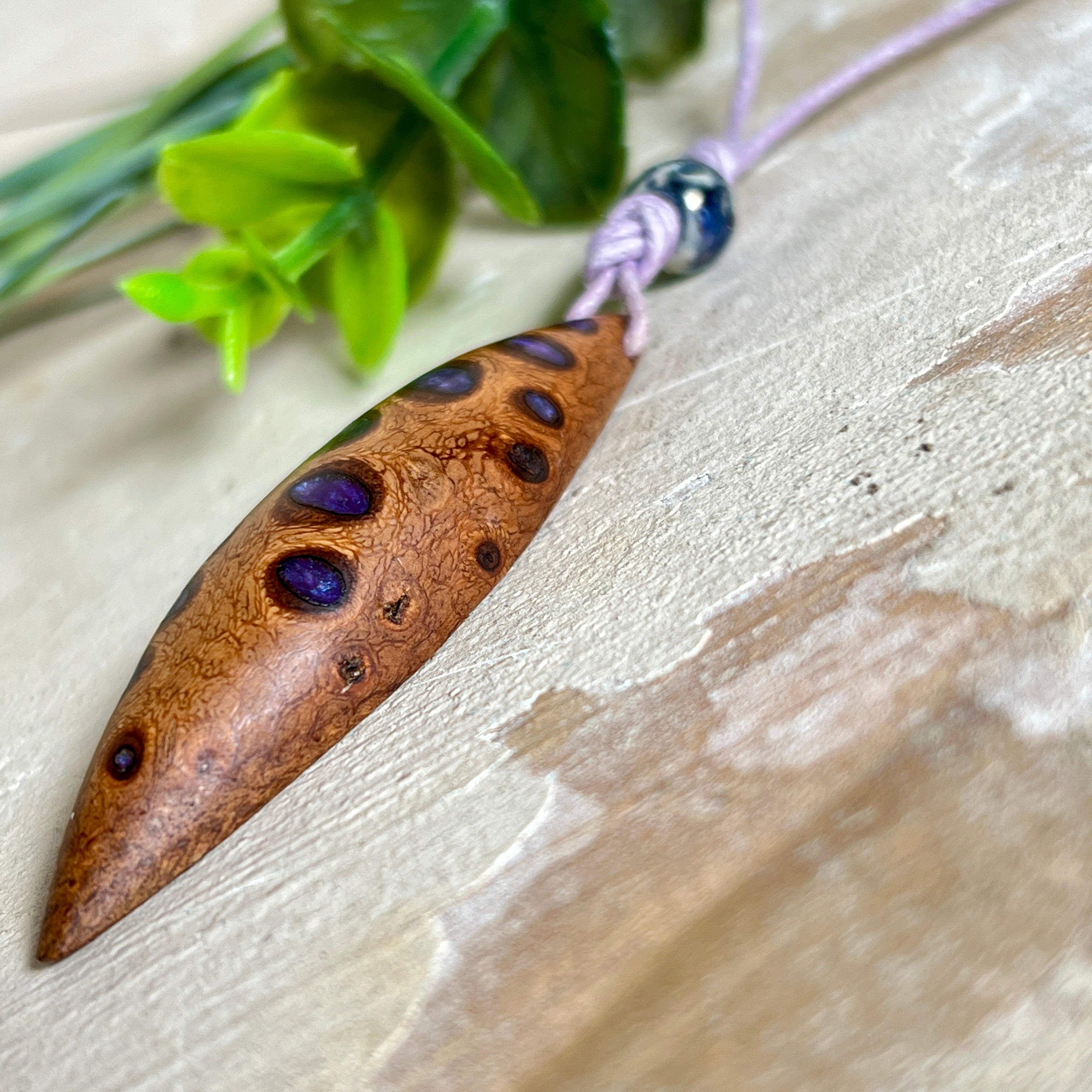 Handgefertigter Anhänger aus einem australischen Banksiazapfen mit eingelassenem metallic-violettem Harz. Der ovale, tropfenförmige Anhänger ist ca. 5,5 cm lang und wird mit einem verstellbaren Baumwollband geliefert. Jedes Stück zeigt die einzigartige natürliche Struktur des Zapfens und schimmert in satten Lilatönen – ein stilvoller Schmuck mit organischem Charakter.
