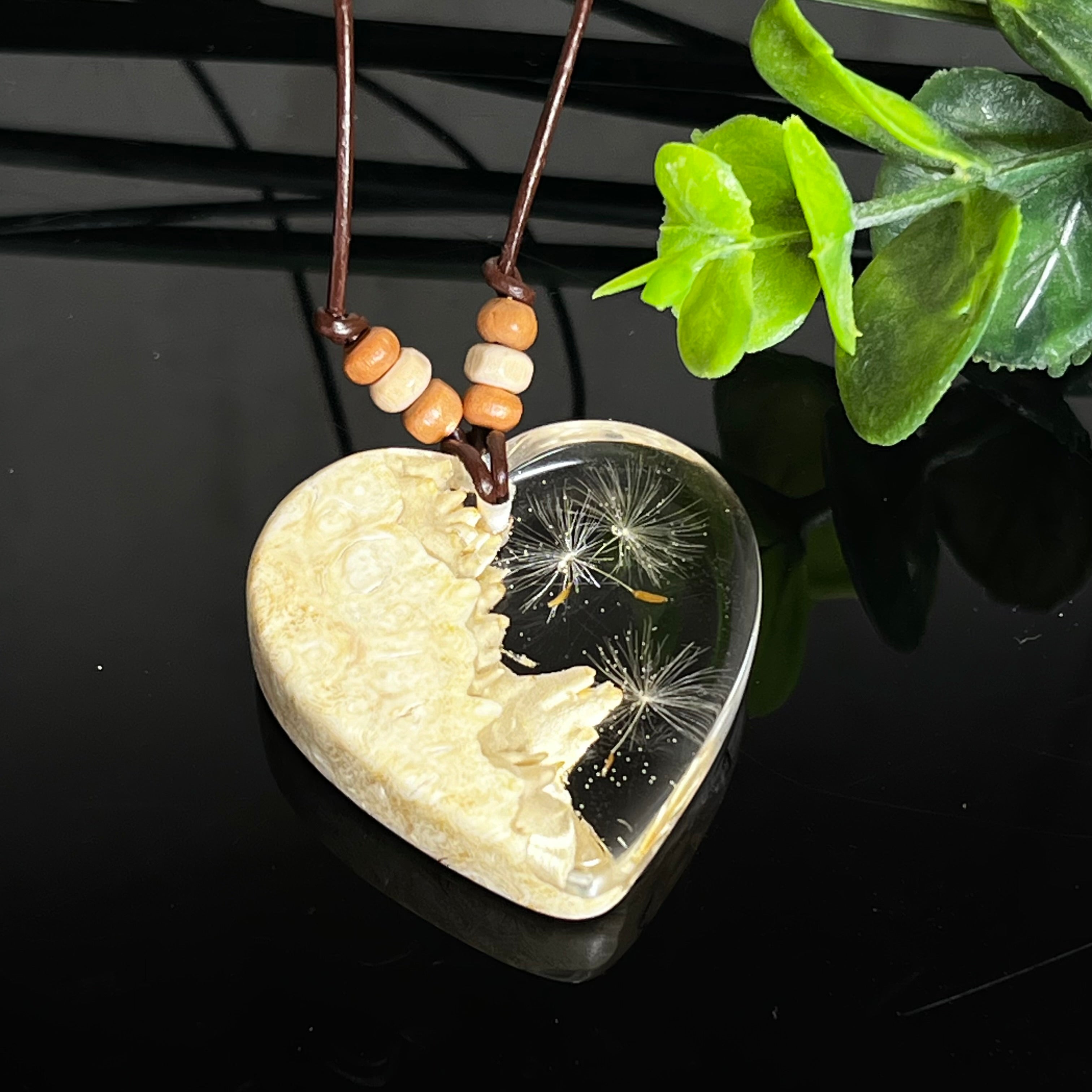 Herzanhänger aus Goldfield-Maserholz und transparentem Epoxidharz mit echter Pusteblume. Das handgefertigte Unikat kombiniert die natürliche Maserung des Holzes mit der zarten Leichtigkeit schwebender Pusteblumenschirmchen. Das braune Lederband mit Holzperlen ist verstellbar und verleiht dem Schmuckstück eine naturnahe, elegante Ausstrahlung.