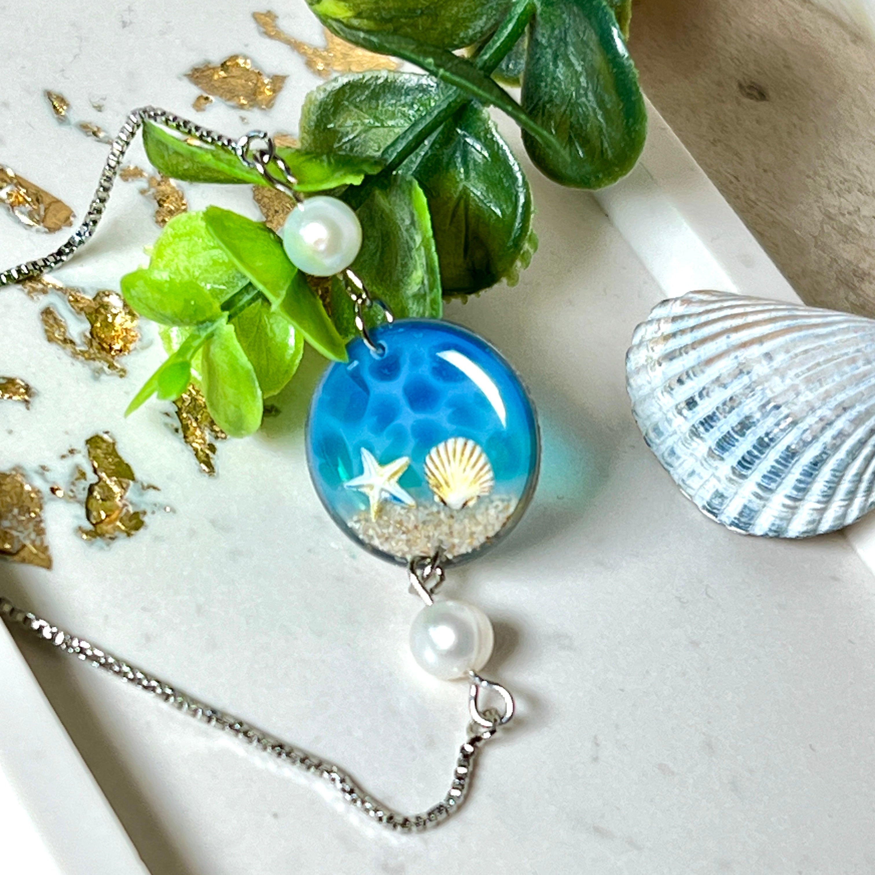 Elegantes Edelstahl-Armband mit rundem Resin-Anhänger in Meeresblau. Im Anhänger liegt echter Meersand als Ufersaum; darüber Sticker-Motive Seestern & Muschel. Zwei feine Süßwasserperlen verzieren das Armband. Verstellbarer Schiebeverschluss . Handgefertigtes Unikat – maritimer Schmuck, der an Wellenrauschen und Strandspaziergänge erinnert.