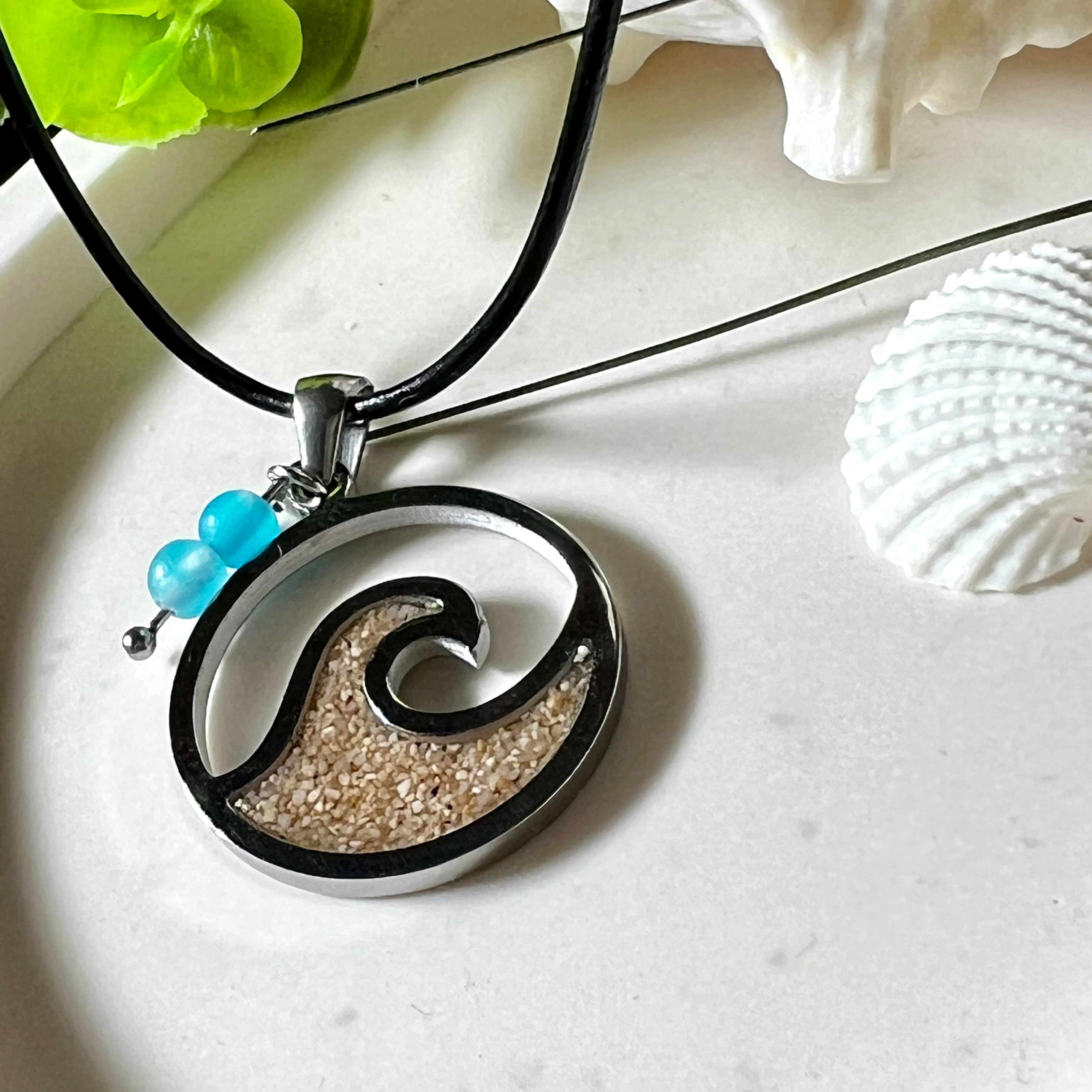 Wave Spirit – runder Edelstahlanhänger mit echter Sandfüllung in Harz eingebettet, inspiriert von einer stilisierten Welle. Verziert mit zwei blauen Achatperlen. Der Anhänger (ca. 2,5 cm Durchmesser) hängt an einer 45 cm langen Lederkette mit Edelstahlverschluss und 5 cm Verlängerungskette. Maritimer Look trifft moderne Eleganz – ein handgefertigtes Schmuckstück für Meerliebhaber