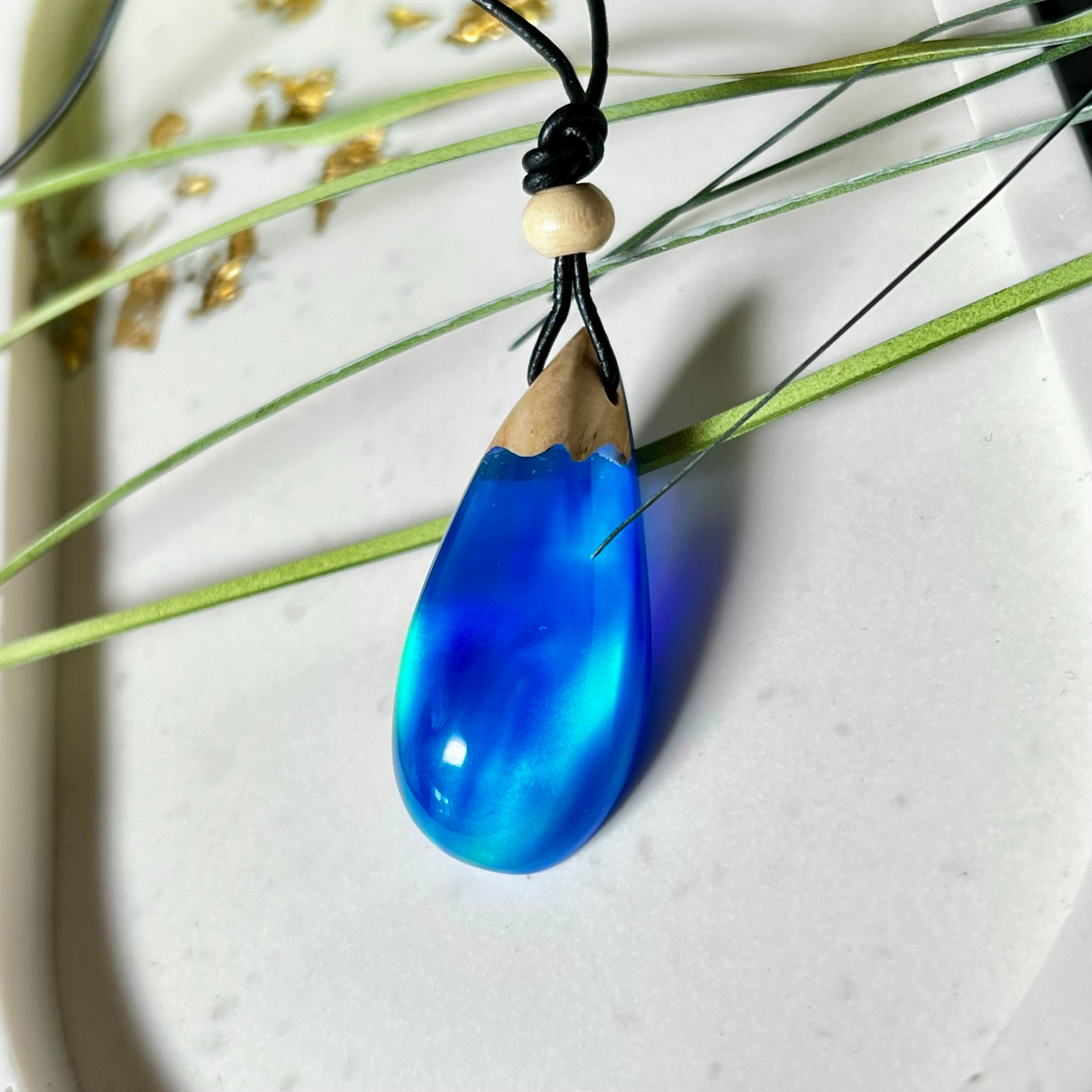 Handgefertigter Tropfenanhänger aus Maserholz und Harz mit synthetischem Opal. Die Farben schimmern je nach Lichteinfall in leuchtendem Blau und Grün – inspiriert von Polarlichtern. Der Anhänger ist ca. 3,9 × 1,8 cm groß und hängt an einem verstellbaren Lederband mit Holzperlen. Jedes Stück ist ein Unikat mit einzigartiger Holzmaserung. Ideal als Geschenk oder stilvoller Begleiter im Alltag.