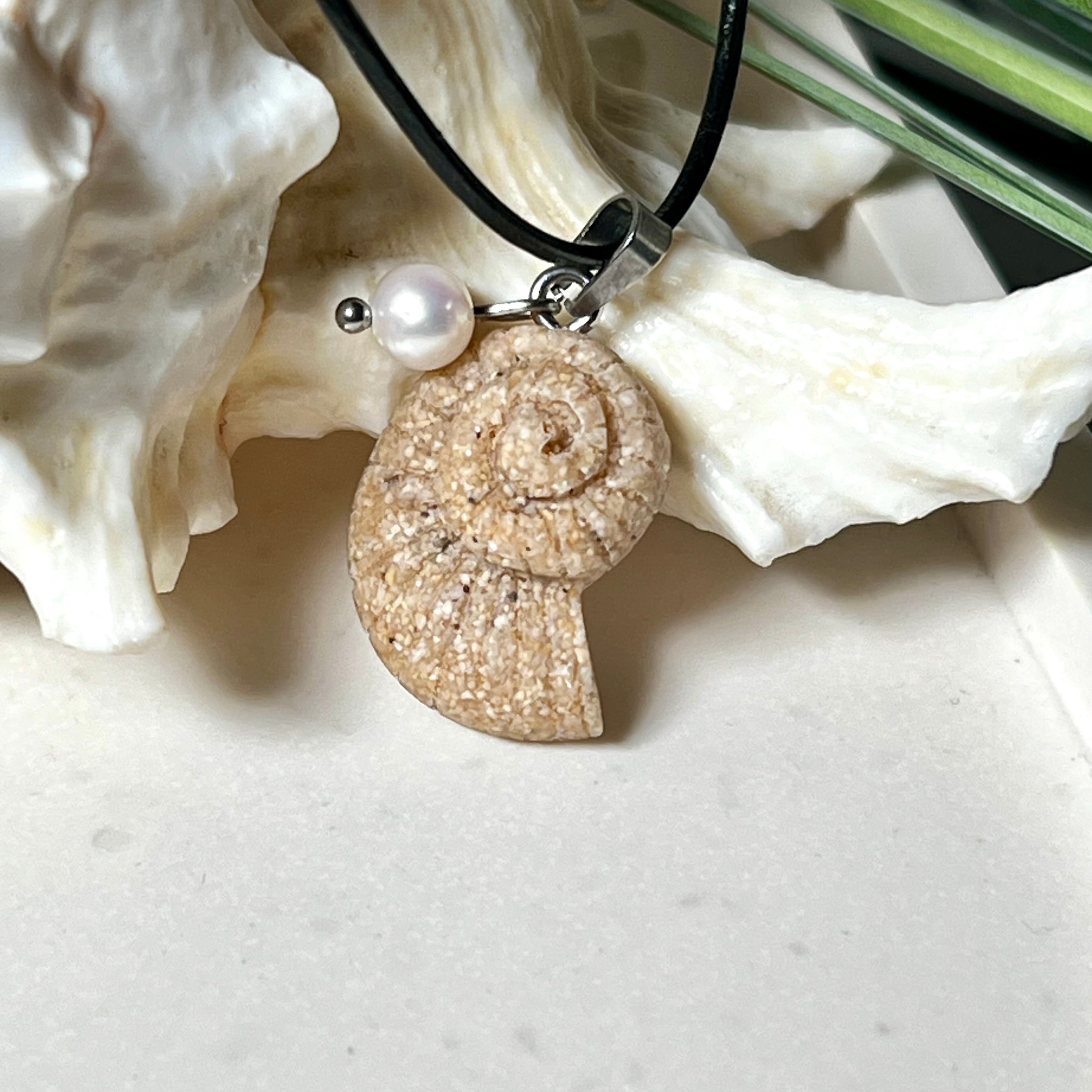 Handgefertigter Anhänger in Ammonit-Form aus echtem Meersand und Harz, kombiniert mit einer weißen Süßwasserperle. Der maritime Anhänger hängt an einer schwarzen Lederkette mit Edelstahlöse und Karabinerverschluss. Im Hintergrund liegt eine weiße Muschel, die das natürliche, strandinspirierte Design des Schmuckstücks unterstreicht. Ideal als besonderes Geschenk oder Erinnerung an den Sommer.