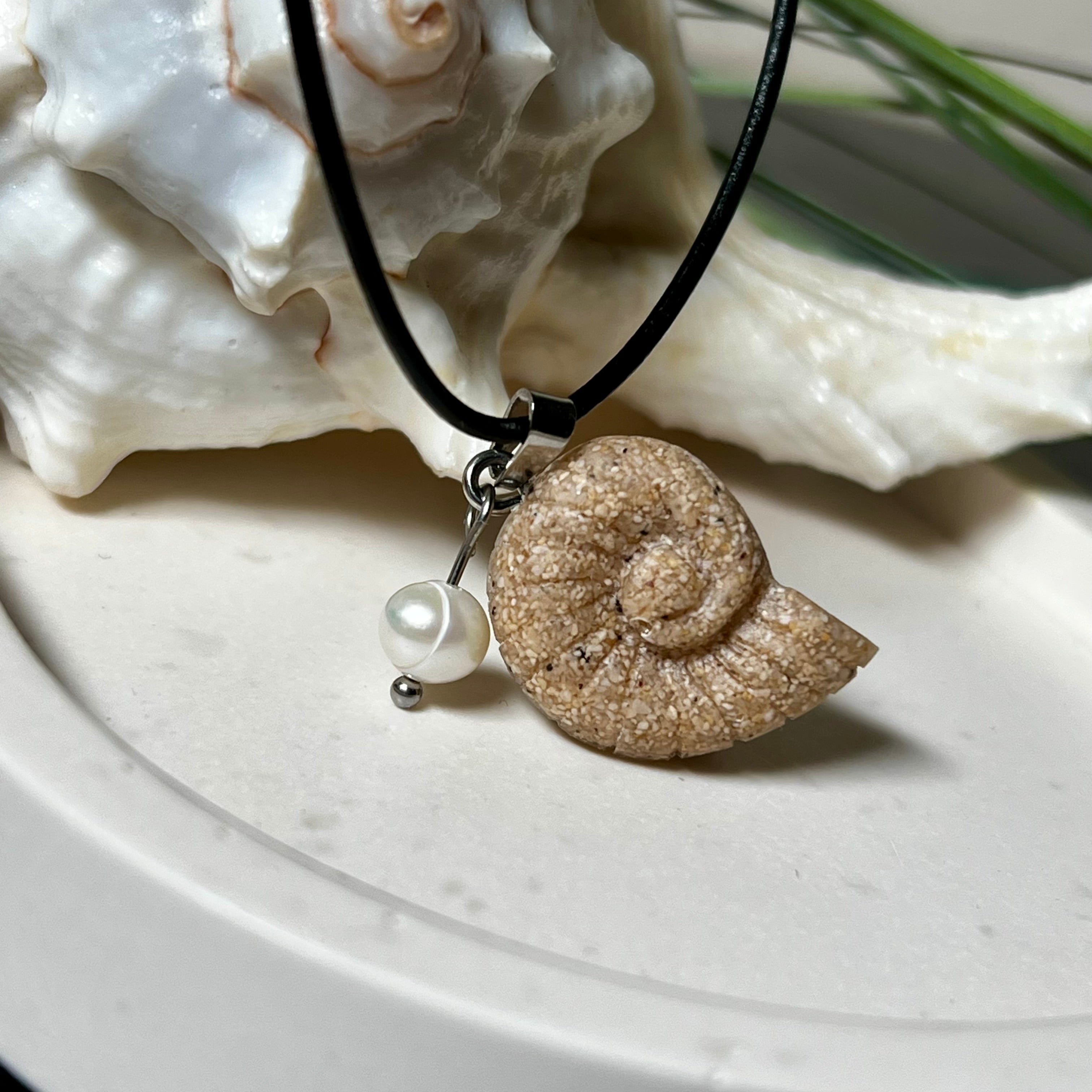 Handgefertigter Ammonit-Anhänger aus echtem Meersand & Harz mit filigraner Edelstahlöse und einer edlen Süßwasserperle. Maritimer Schmuck, inspiriert von der Kraft und Schönheit des Ozeans. Jedes Stück ein Unikat. Präsentiert auf einer Muschel, mit Lederkette.