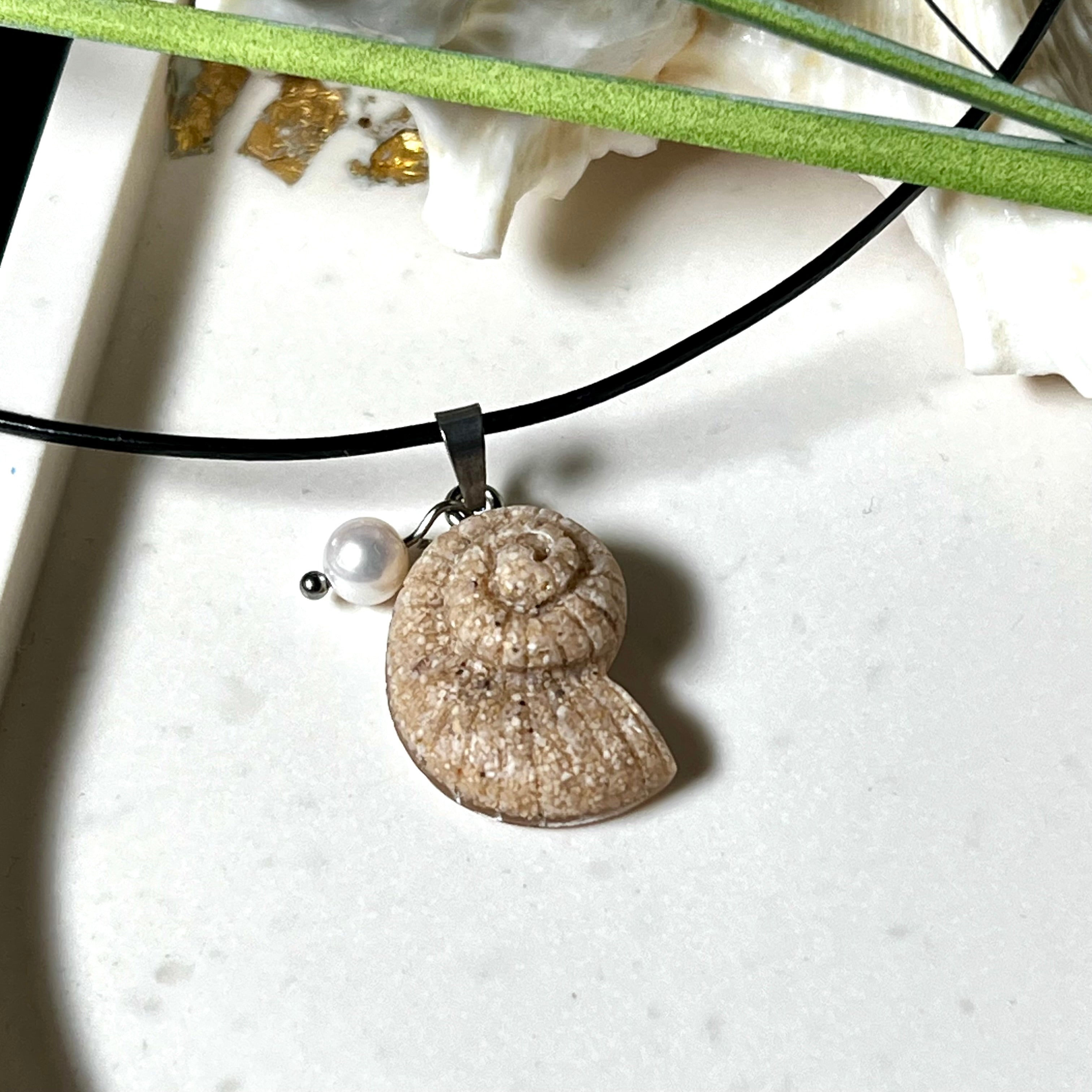 Handgefertigter Anhänger in Ammonit-Form aus echtem Meersand und Harz, kombiniert mit einer weißen Süßwasserperle. Der maritime Anhänger hängt an einer schwarzen Lederkette mit Edelstahlöse und Karabinerverschluss. Im Hintergrund liegt eine weiße Muschel, die das natürliche, strandinspirierte Design des Schmuckstücks unterstreicht. Ideal als besonderes Geschenk oder Erinnerung an den Sommer.