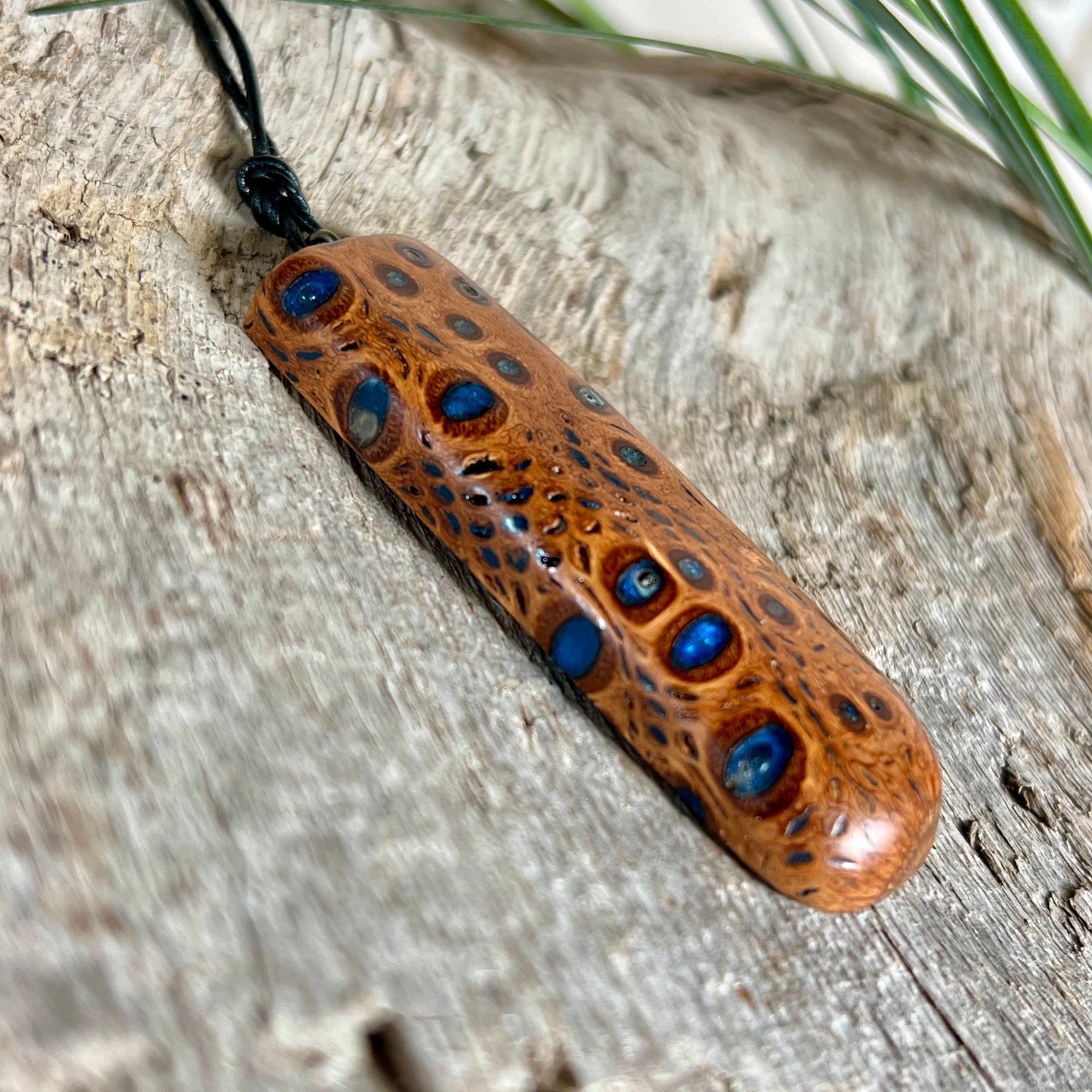 Handgefertigter Anhänger aus australischem Banksiazapfen und gegossenem Harz in Blau. Der rechteckige Anhänger (5,5 x 2 cm) zeigt eine charakteristische Maserung mit markanten Löchern, die durch die Samenstruktur des Zapfens entstehen. Das Lederband ist verstellbar (29–51 cm) und mit Holzperlen verziert. Ein edles Natur-Unikat.
