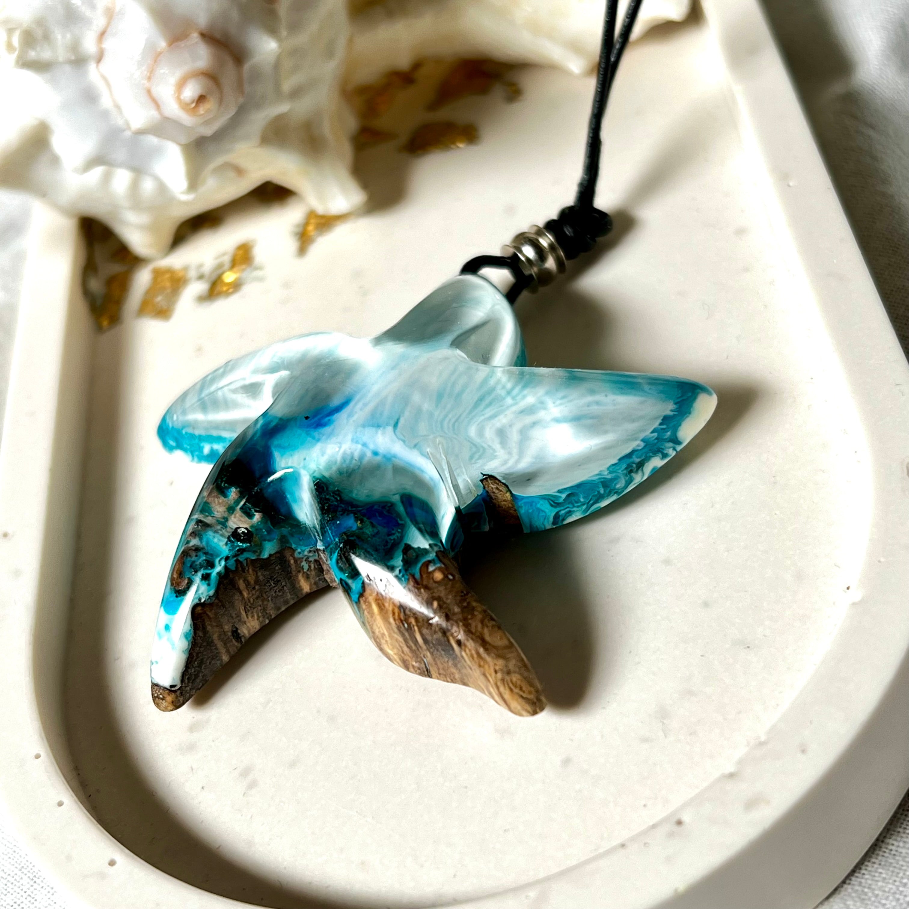 Handgefertigte Halskette in Seestern-Form mit Anhänger aus Naturholz und blau-weiß marmoriertem Epoxidharz. Das Schmuckstück ist einzigartig gestaltet und spiegelt maritimen Stil und Naturverbundenheit wider. Getragen an einem verstellbaren Lederband mit Holzperlen. Ideal für Meeresliebhaber und Naturfreunde.