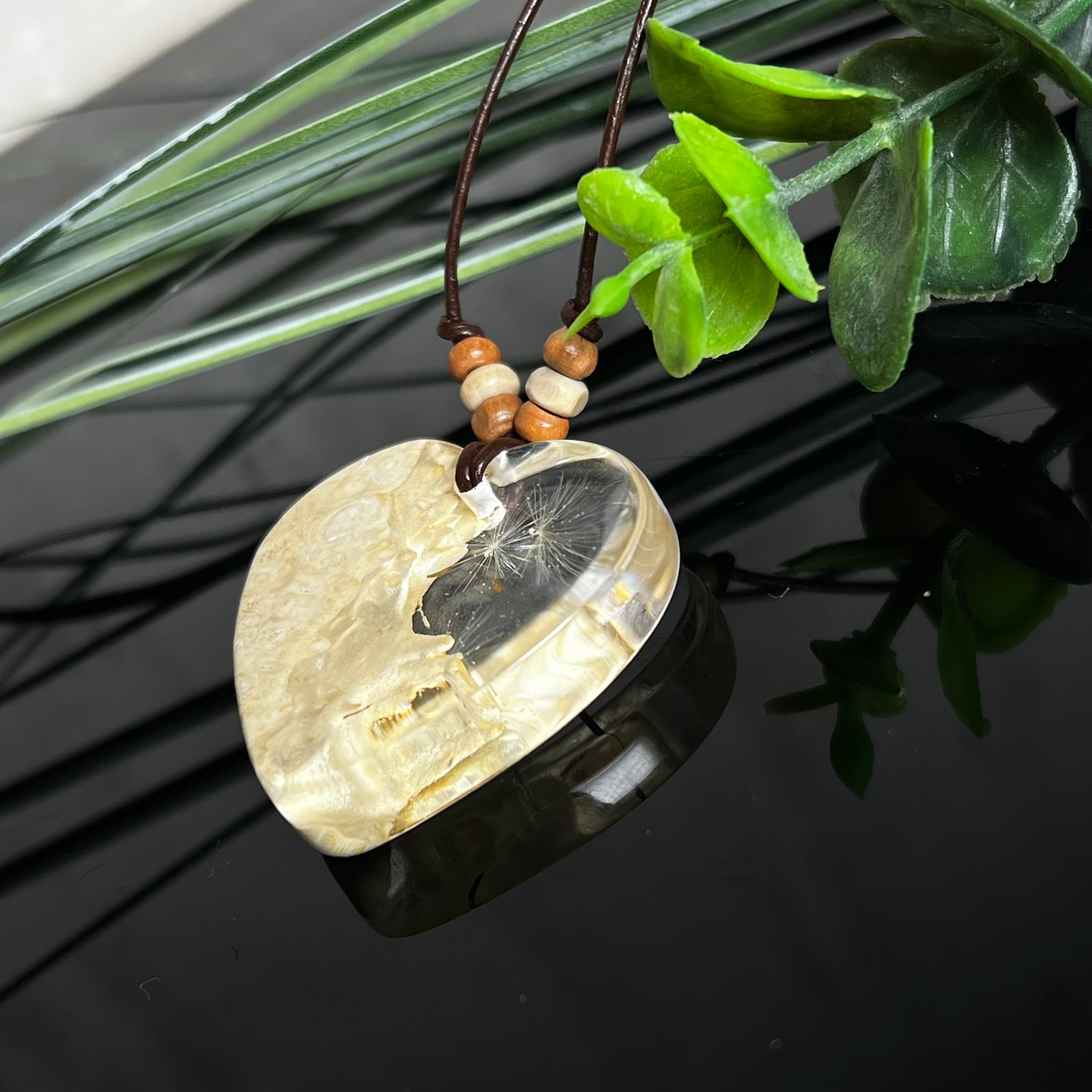 Herzanhänger aus Goldfield-Maserholz und transparentem Epoxidharz mit echter Pusteblume. Das handgefertigte Unikat kombiniert die natürliche Maserung des Holzes mit der zarten Leichtigkeit schwebender Pusteblumenschirmchen. Das braune Lederband mit Holzperlen ist verstellbar und verleiht dem Schmuckstück eine naturnahe, elegante Ausstrahlung.
