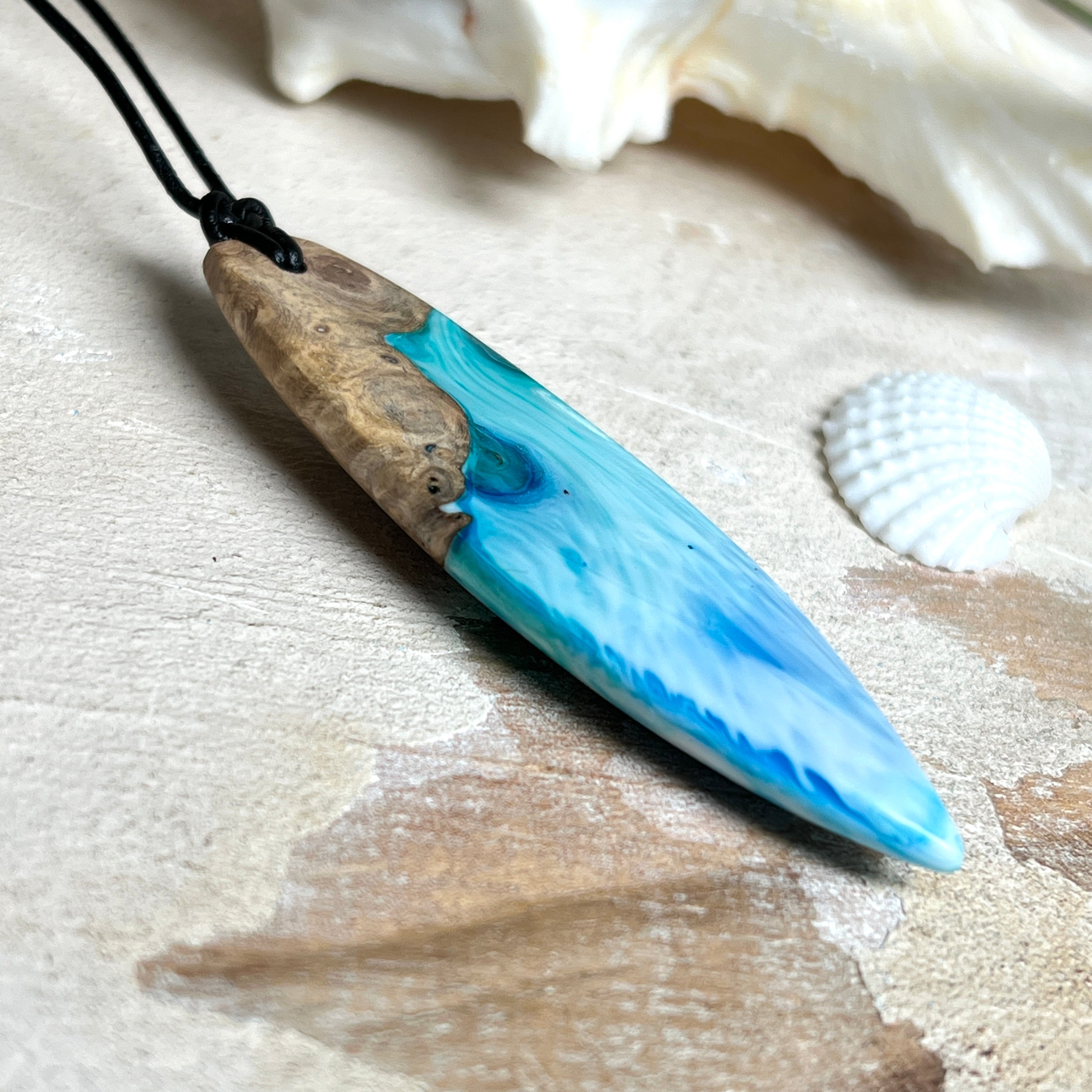 Surfbrett-Anhänger handgefertigt aus Maserholz & Harz in Blau-Weiß. Größe ca. 6,3 × 2 cm. Lederband verstellbar 26–47 cm, Schiebeknoten mit Holzperlen.