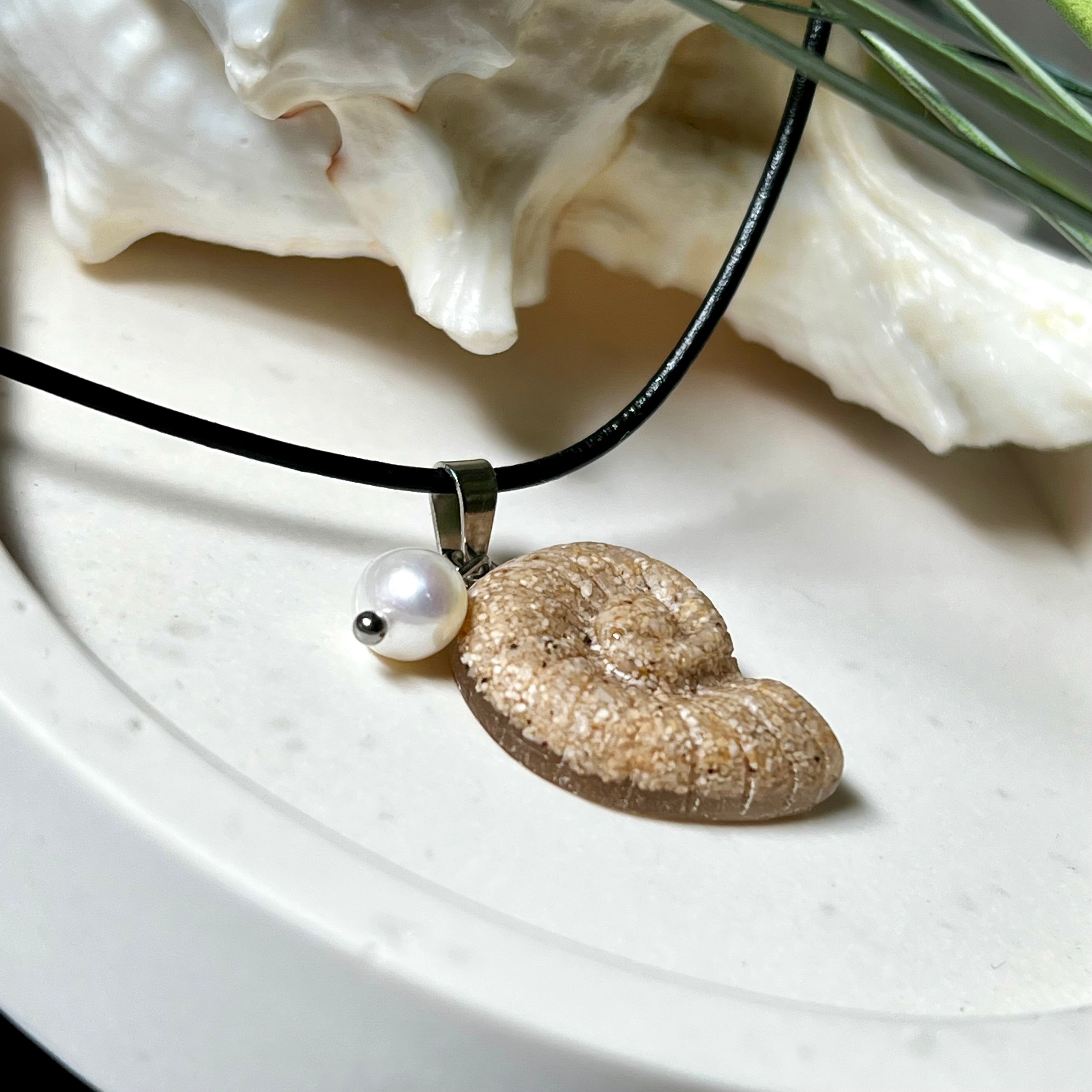Handgefertigter Ammonit-Anhänger aus echtem Meersand & Harz mit filigraner Edelstahlöse und einer edlen Süßwasserperle. Maritimer Schmuck, inspiriert von der Kraft und Schönheit des Ozeans. Jedes Stück ein Unikat. Präsentiert auf einer Muschel, mit Lederkette.