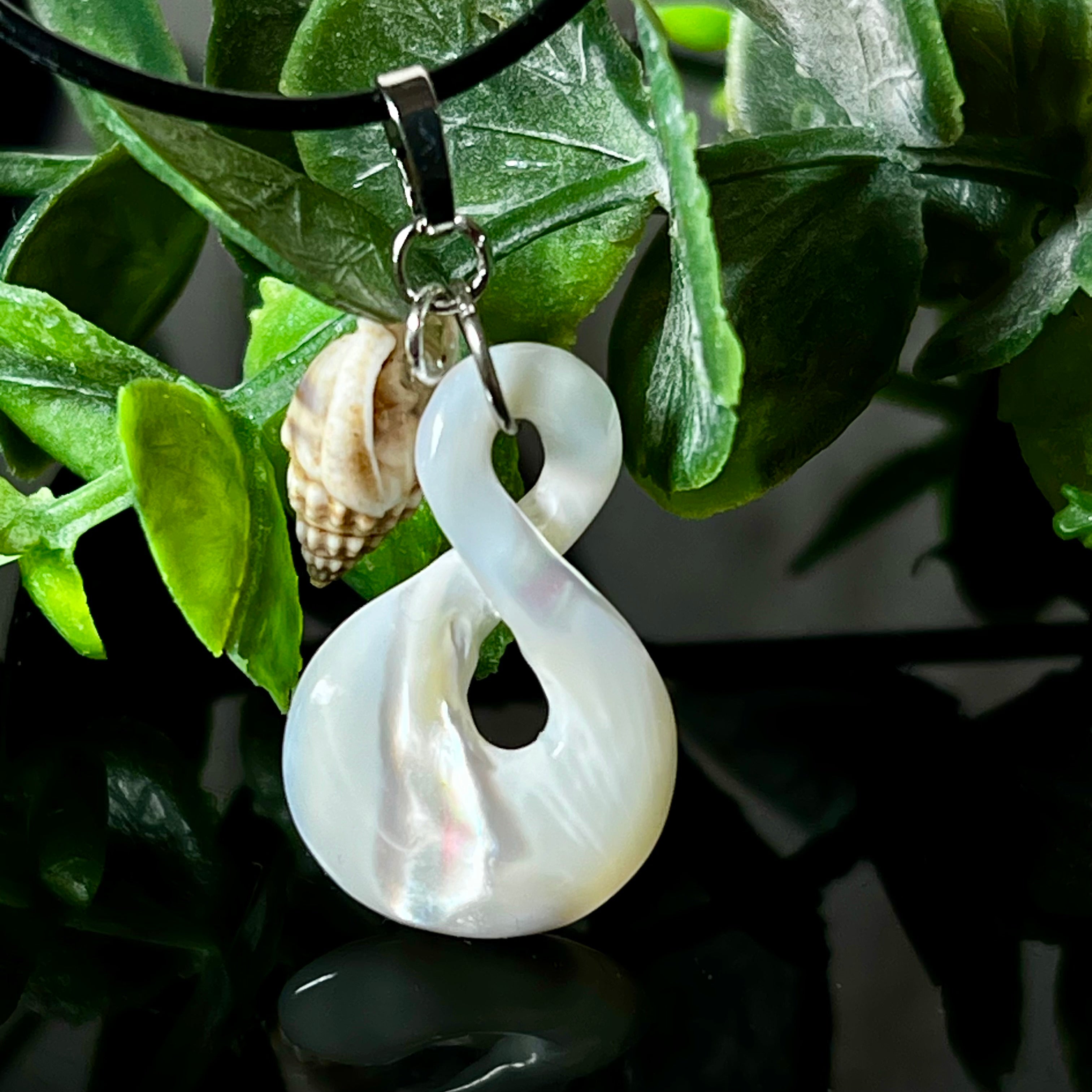 Detailaufnahme eines handgefertigten Anhängers aus echtem Perlmutt in Schlaufenform, kombiniert mit einer kleinen echten Muschel als Charm-Anhänger. Der Schmuck hängt an einer feinen schwarzen Lederschnur mit Edelstahlverschluss und symbolisiert Harmonie und Unendlichkeit. Ideal für Meeresliebhaberinnen und Fans von Naturmaterialien.