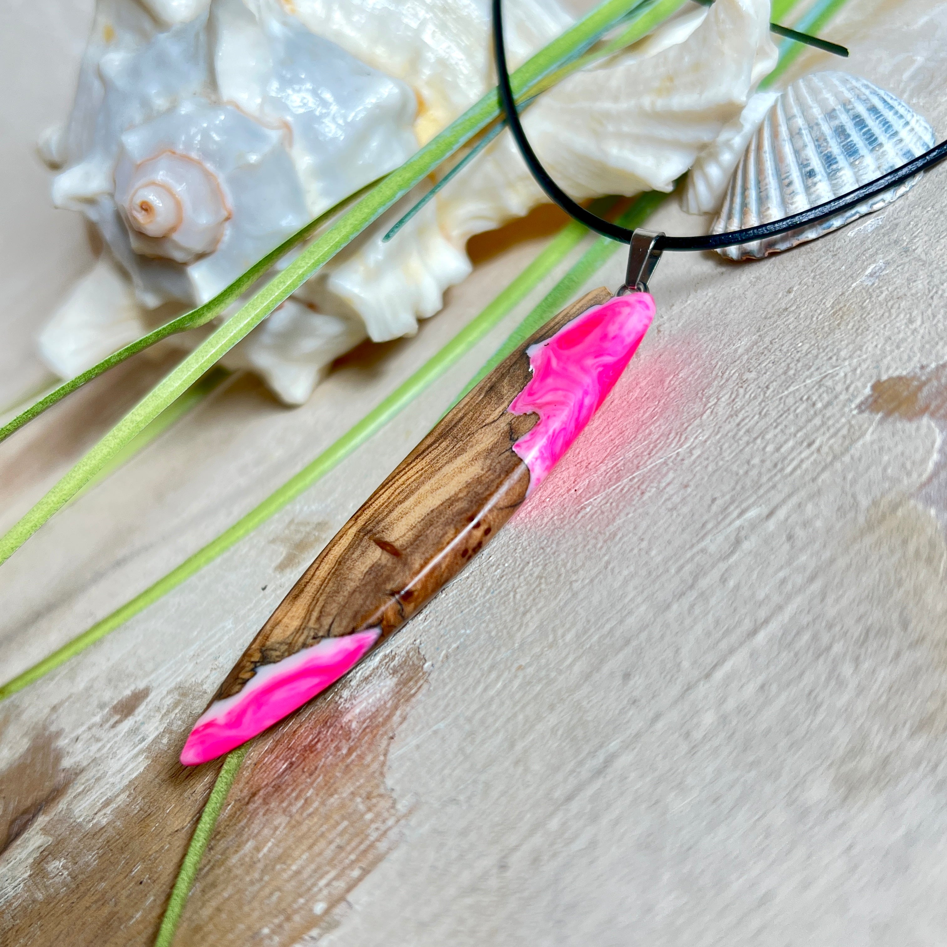 Handgefertigter Surfbrett-Anhänger „Pink Glow“ aus edlem Olivenholz und fluoreszierendem Epoxidharz in Pink & Weiß. Fluoreszierend bei Schwarzlicht, mit Edelstahl-Anhängerschlaufe und schwarzer Lederkette (45 cm + 5 cm Verlängerung). Ein maritimes Unikat – perfekt als Geschenk für Surfer und Meeresliebhaber.