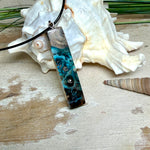 Rechteckiger Stabanhänger aus Maserholz und glasklarem Epoxidharz in tiefen Blau- und Türkistönen mit organischer Lagunen/Riff-Optik. Hochglanzpoliert, ca. 5,3 × 1,2 cm. Der Anhänger hängt an einer schwarzen Lederkette mit Edelstahl-Karabiner und 5-cm-Verlängerung (45 + 5 cm); die Anhängerschlaufe ist ebenfalls aus Edelstahl. Handgefertigtes Unikat, Lieferung in edler Schmuckbox – maritimer Look.