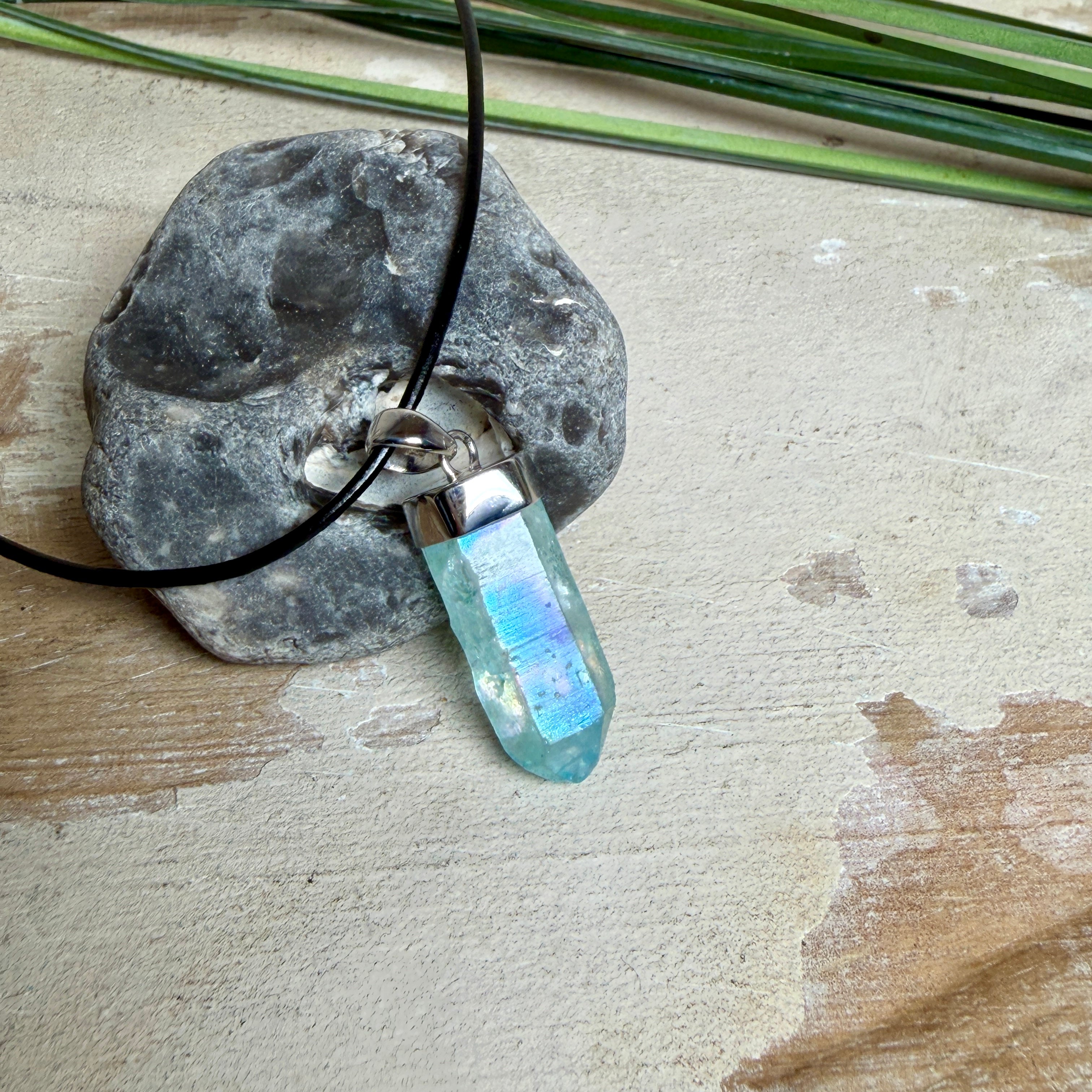 Nahaufnahme einer handgefertigten Halskette mit Aqua Aura Rohkristall: naturbelassener, sechskantiger Bergkristall mit goldbedampfter Aqua-Beschichtung in Türkisblau, gefasst in 925er Silber und getragen an einer schwarzen Lederkette mit Edelstahl-Karabiner und Verlängerungskettchen.