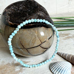 Zartes Larimar-Armband „Lagunenlicht“ mit facettierten 3 mm Perlen aus seltenem Atlantisstein auf Elastikfaden. Die kleinen Larimarsteine zeigen sanfte Blau- und Weißtöne wie Lagunenwasser. Innenumfang ca. 19 cm. Fotografiert auf Muscheln, einem dekorativen Stein und in einer schwarzen Schmuckbox von ZeitlosSchmuckDesign im maritimen Stil.
