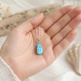 Filigrane Larimar-Halskette aus 925 Silber auf einer Frauenhand, mit elegantem maritimem Hintergrund – handgefertigter Natursteinschmuck in sanftem Karibikblau, edle Damenkette mit tropfenförmigem Larimar-Anhänger.