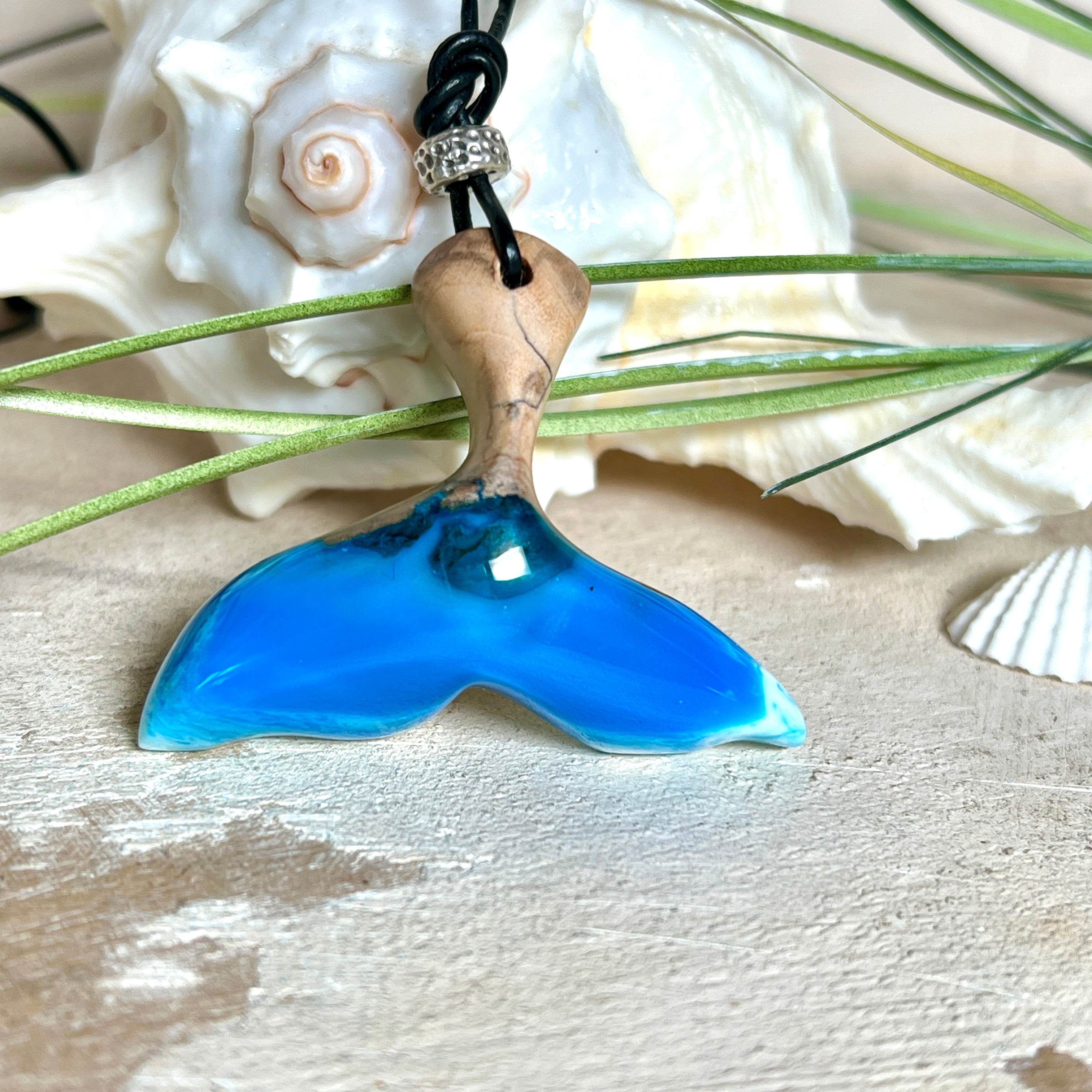 Handgefertigter Walflossen-Anhänger aus Olivenholz und glänzendem Harz in Blau-Grün-Weiß – maritimer Schmuck „Flossenklang“. Der Anhänger erinnert an die Form einer Walflosse und vereint die natürliche Maserung des Holzes mit dem schimmernden Farbspiel des Harzes. Mit verstellbarem schwarzen Lederband und Spacerperle. Ideal für Meeresliebhaber und Naturliebhaber – jedes Stück ein Unikat.