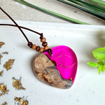 Handgefertigter Herzanhänger aus Olivenholz und leuchtend pinkem Epoxidharz mit eingeschlossenen Pusteblumensamen. Verstellbares braunes Lederband mit Holzperlen, Anhänger ca. 3,5 × 3,3 cm groß. Das Harz leuchtet dezent unter Schwarzlicht. Ein einzigartiges Unikat von ZeitlosSchmuckDesign, inspiriert von Kindheitsglück und Pusteblumen.