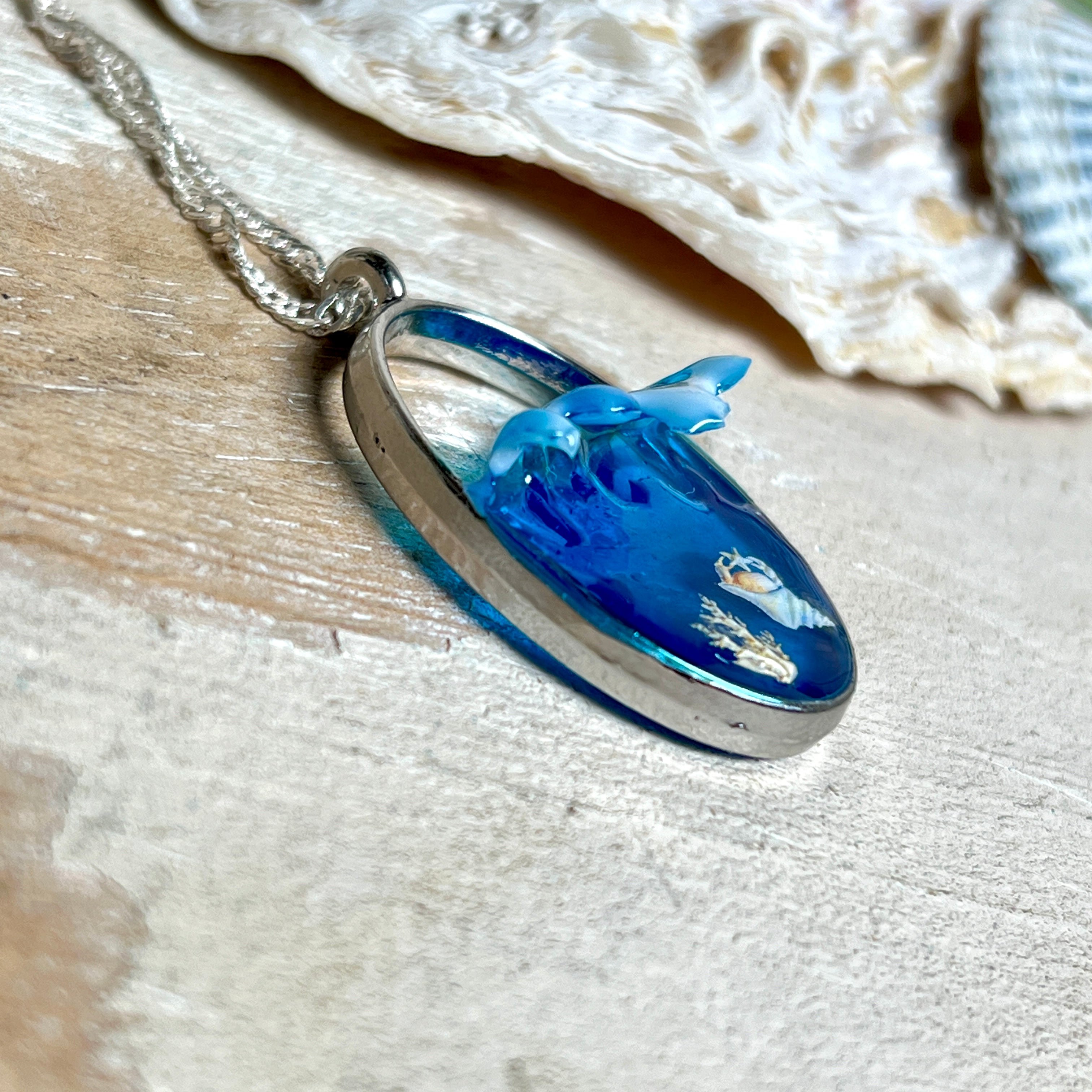 „Riffschatz“ – ovaler Harz-Anhänger mit plastischer 3D-Welle in Blau/Türkis, weißer Schaumkrone und Muschel-/Korallen-Stickern am Grund. Die Sticker sind unter klarem Harz versiegelt. Silber-Legierung, ca. 3,9 × 2,3 cm, an einer ca. 56 cm langen versilberten Kette. Handgefertigt, hochglänzend, jedes Stück ein Unikat.