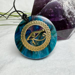 Runder Anhänger mit goldenem Horusauge aus Metall in blau türkis violettem Epoxidharz am schwarzen Baumwollband, spiritueller Eye of Ra Naturschmuck Amulett