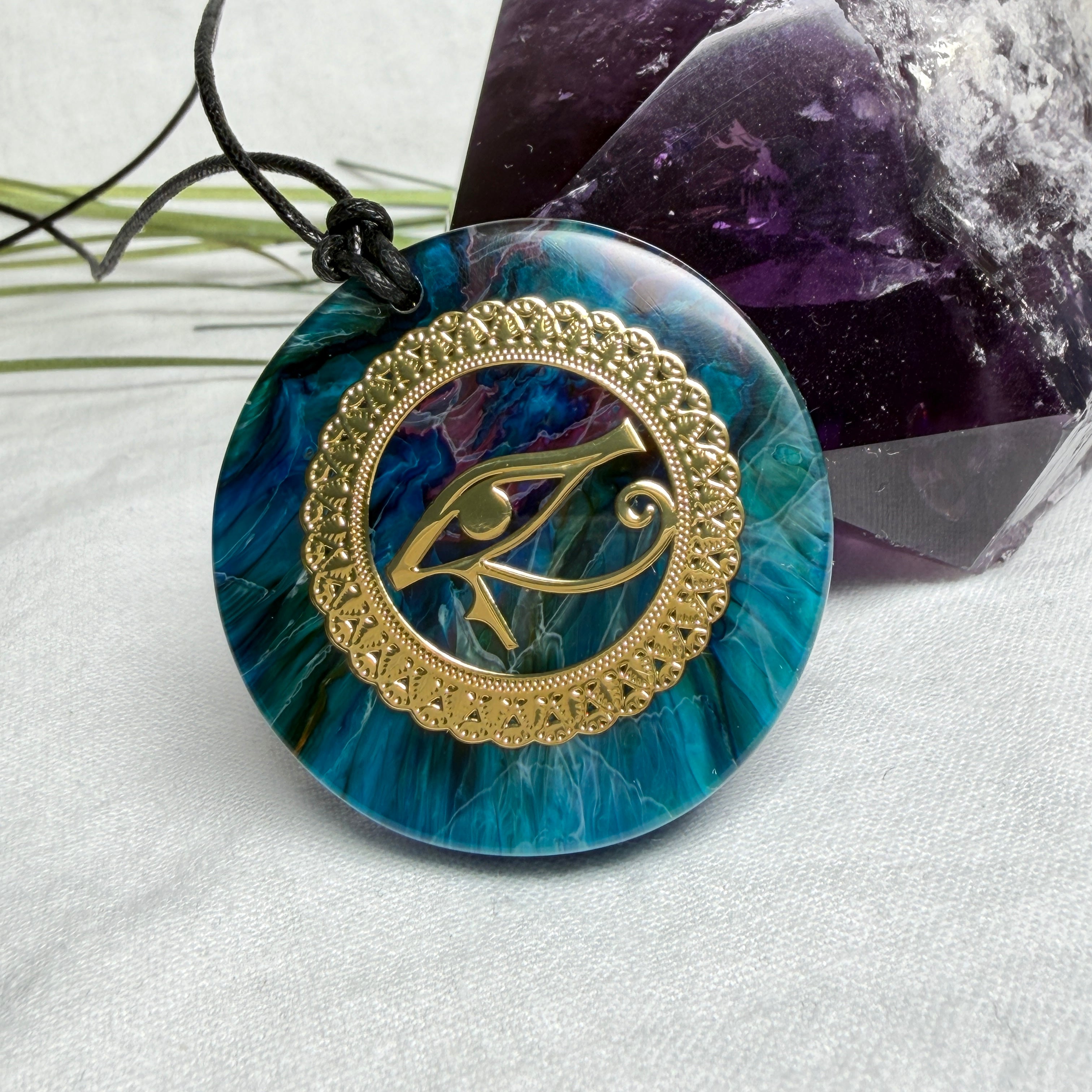 Runder Anhänger mit goldenem Horusauge aus Metall in blau türkis violettem Epoxidharz am schwarzen Baumwollband, spiritueller Eye of Ra Naturschmuck Amulett