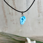 Larimar Freeform-Anhänger auf schwarzer Lederkette. Beidseitig tragbarer Trommelstein, ca. 2 x 1,8 cm groß, mit Silberöse. Der seltene, kostbare Larimar zeigt lebendige Blau- und Weißnuancen wie karibisches Lagunenwasser. Lederkette 45 cm mit Edelstahl-Karabiner und 5 cm Edelstahl-Verlängerung, fotografiert vor einer großen Muschel im maritimen Stil.