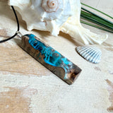 Rechteckiger Stabanhänger aus Maserholz und glasklarem Epoxidharz in tiefen Blau- und Türkistönen mit organischer Lagunen/Riff-Optik. Hochglanzpoliert, ca. 5,3 × 1,2 cm. Der Anhänger hängt an einer schwarzen Lederkette mit Edelstahl-Karabiner und 5-cm-Verlängerung (45 + 5 cm); die Anhängerschlaufe ist ebenfalls aus Edelstahl. Handgefertigtes Unikat, Lieferung in edler Schmuckbox – maritimer Look.