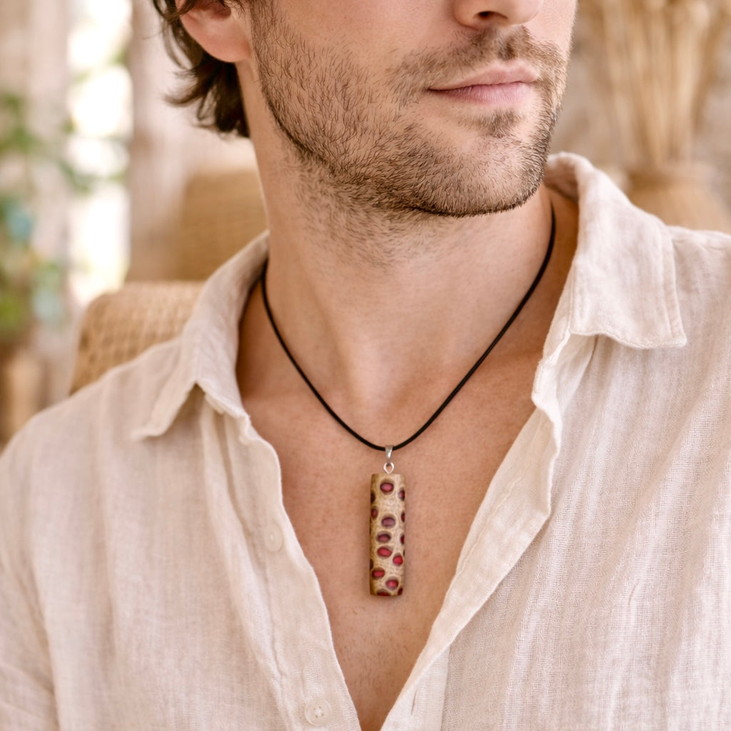 Handgefertigter Halsanhänger aus echtem Banksiazapfen, veredelt mit rotem Harz in den natürlichen Kammern; ca. 5,5 × 1,6 cm. Mit Anhängerschlaufe aus Edelstahl an schwarzer Lederkette (45 cm + 5 cm Verlängerung) mit Edelstahl-Karabiner. Detailaufnahme bzw. Tragebild vor neutralem, natürlichem Hintergrund.