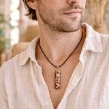 Handgefertigter Halsanhänger aus echtem Banksiazapfen, veredelt mit rotem Harz in den natürlichen Kammern; ca. 5,5 × 1,6 cm. Mit Anhängerschlaufe aus Edelstahl an schwarzer Lederkette (45 cm + 5 cm Verlängerung) mit Edelstahl-Karabiner. Detailaufnahme bzw. Tragebild vor neutralem, natürlichem Hintergrund.