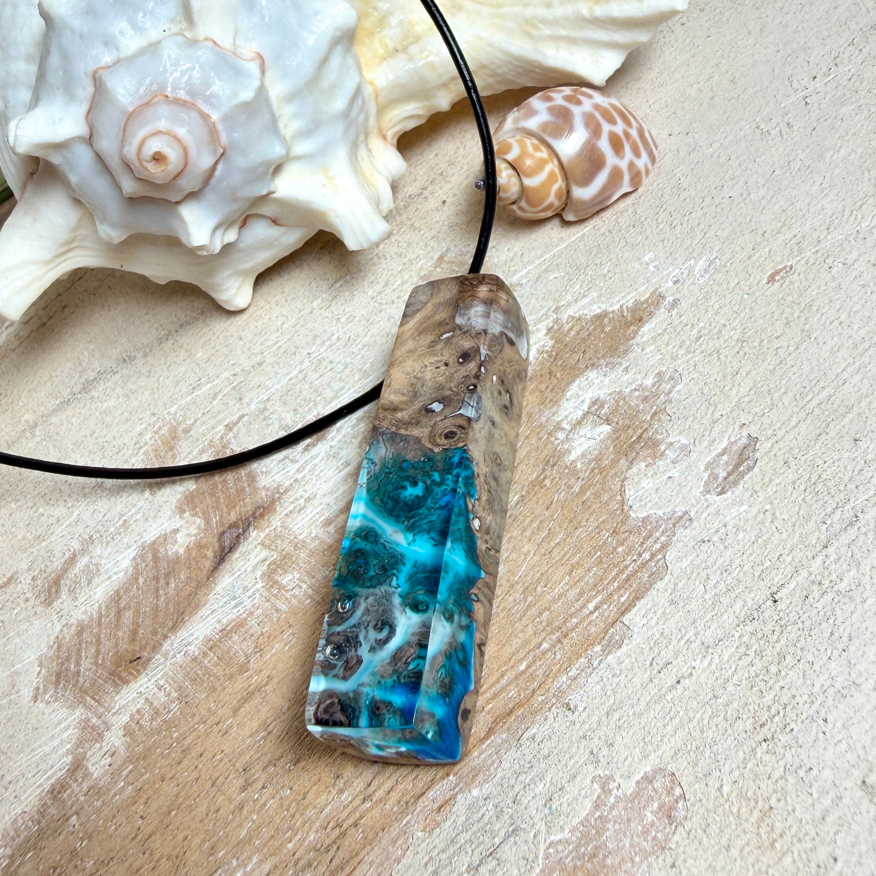 Rechteckiger Stabanhänger aus Maserholz und glasklarem Epoxidharz in tiefen Blau- und Türkistönen mit organischer Lagunen/Riff-Optik. Hochglanzpoliert, ca. 5,3 × 1,2 cm. Der Anhänger hängt an einer schwarzen Lederkette mit Edelstahl-Karabiner und 5-cm-Verlängerung (45 + 5 cm); die Anhängerschlaufe ist ebenfalls aus Edelstahl. Handgefertigtes Unikat, Lieferung in edler Schmuckbox – maritimer Look.