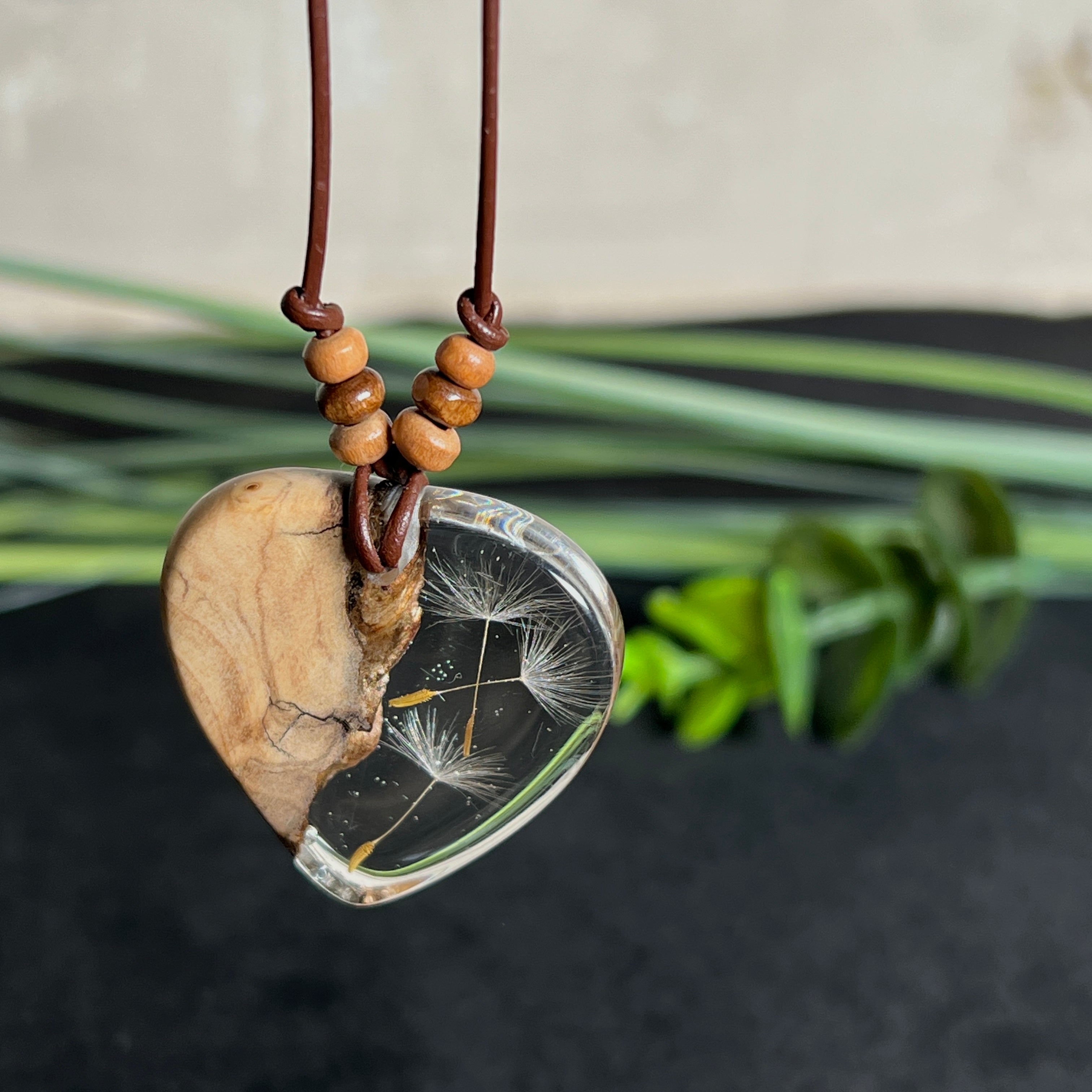 Handgefertigter Herzanhänger aus Olivenholz und klarem Harz mit echten Pusteblumen. Der Anhänger misst ca. 3,5 cm, das Lederband ist verstellbar von 23–43 cm und mit kleinen Holzperlen verziert. Ein natürliches Unikat, Symbol für Wünsche und Glück – sorgfältig gegossen, gesägt, geschliffen und poliert.