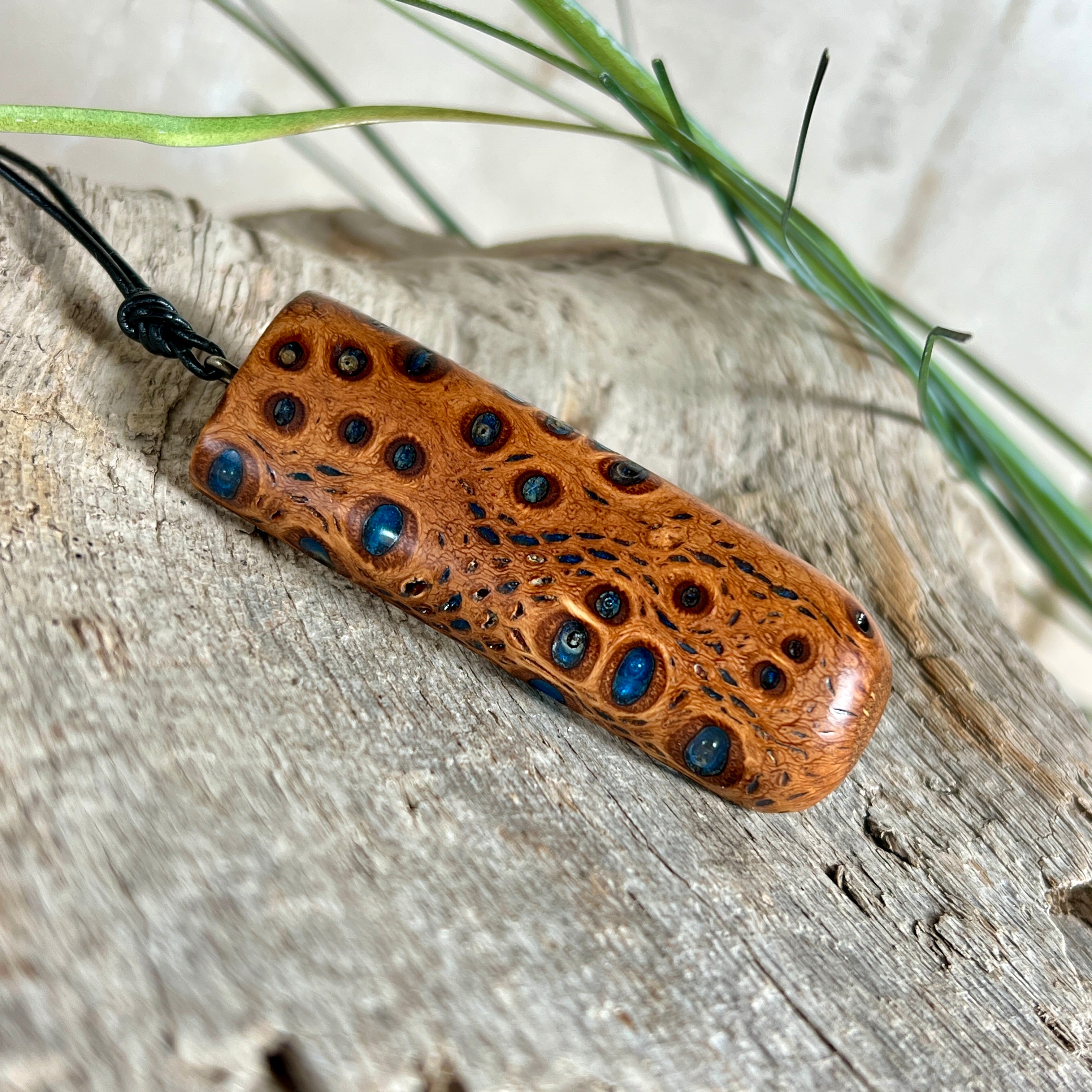 Handgefertigter Anhänger aus australischem Banksiazapfen und gegossenem Harz in Blau. Der rechteckige Anhänger (5,5 x 2 cm) zeigt eine charakteristische Maserung mit markanten Löchern, die durch die Samenstruktur des Zapfens entstehen. Das Lederband ist verstellbar (29–51 cm) und mit Holzperlen verziert. Ein edles Natur-Unikat.